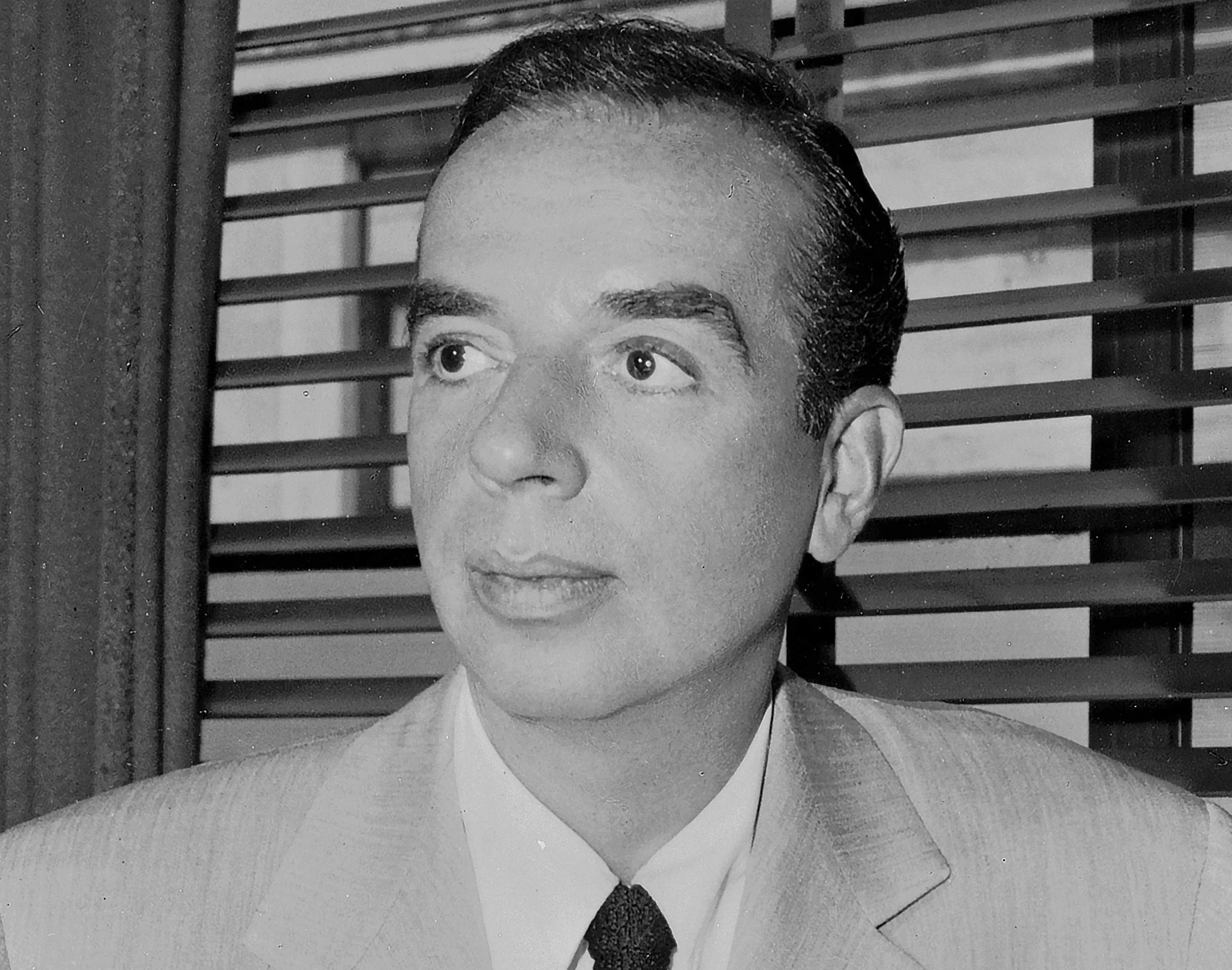 Black and white photograph of a man with slicked-back dark hair, wearing a light-colored suit, white shirt, and dark tie, in front of a window with horizontal blinds.