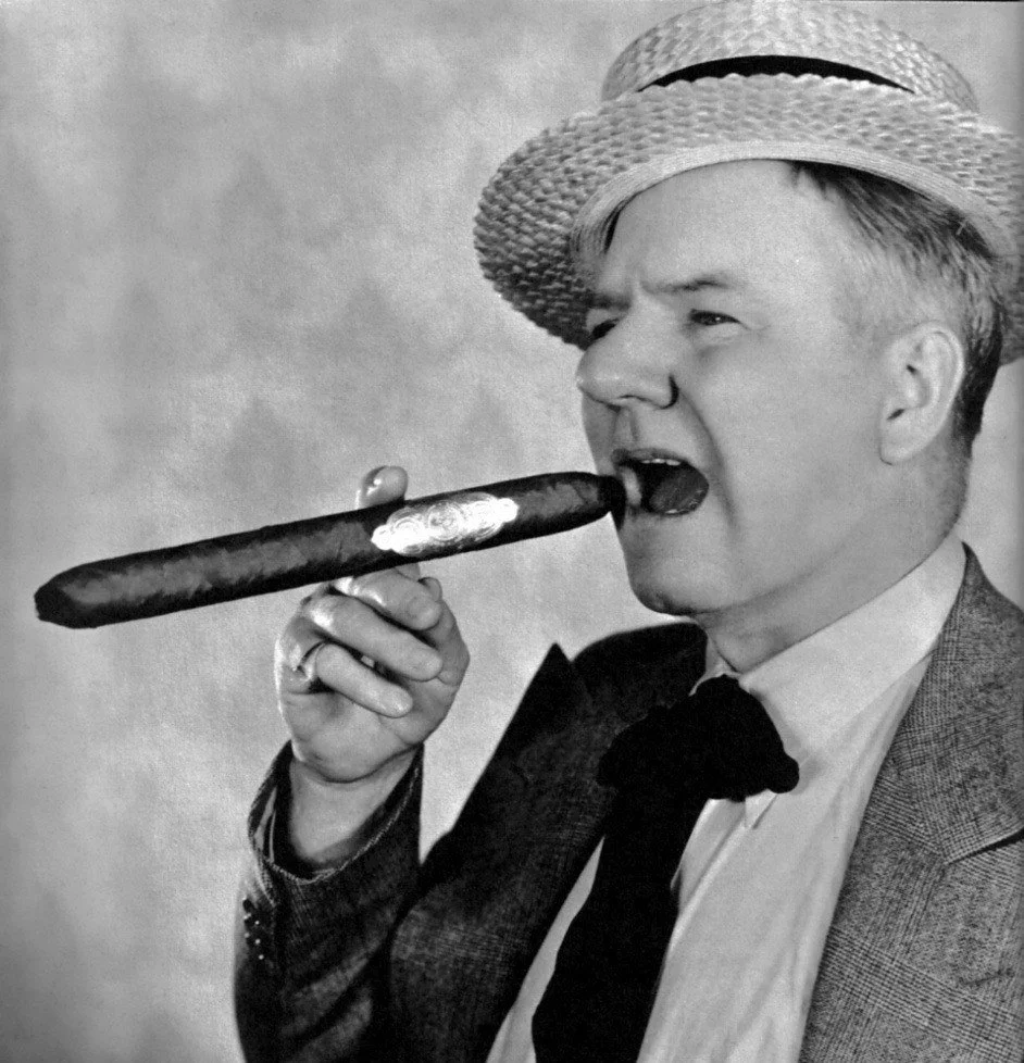 A black and white photo of a man in a suit and tie wearing a straw hat, holding a long cigar in his mouth, and appearing to be biting it.