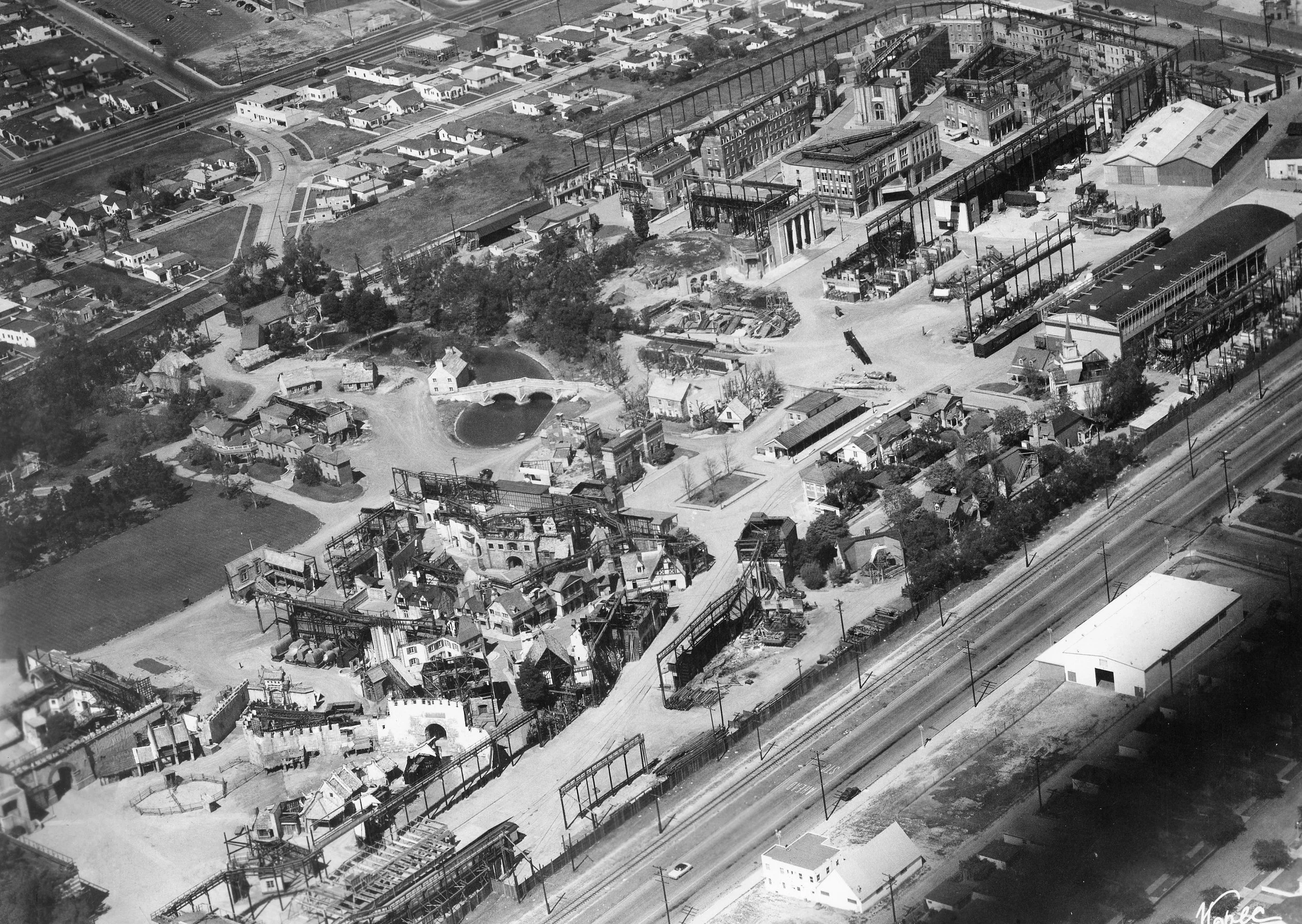 Black and white aerial photograph of a cityscape showing residential houses, a pond with a small bridge, and a large industrial or factory complex with multiple buildings and railway tracks.