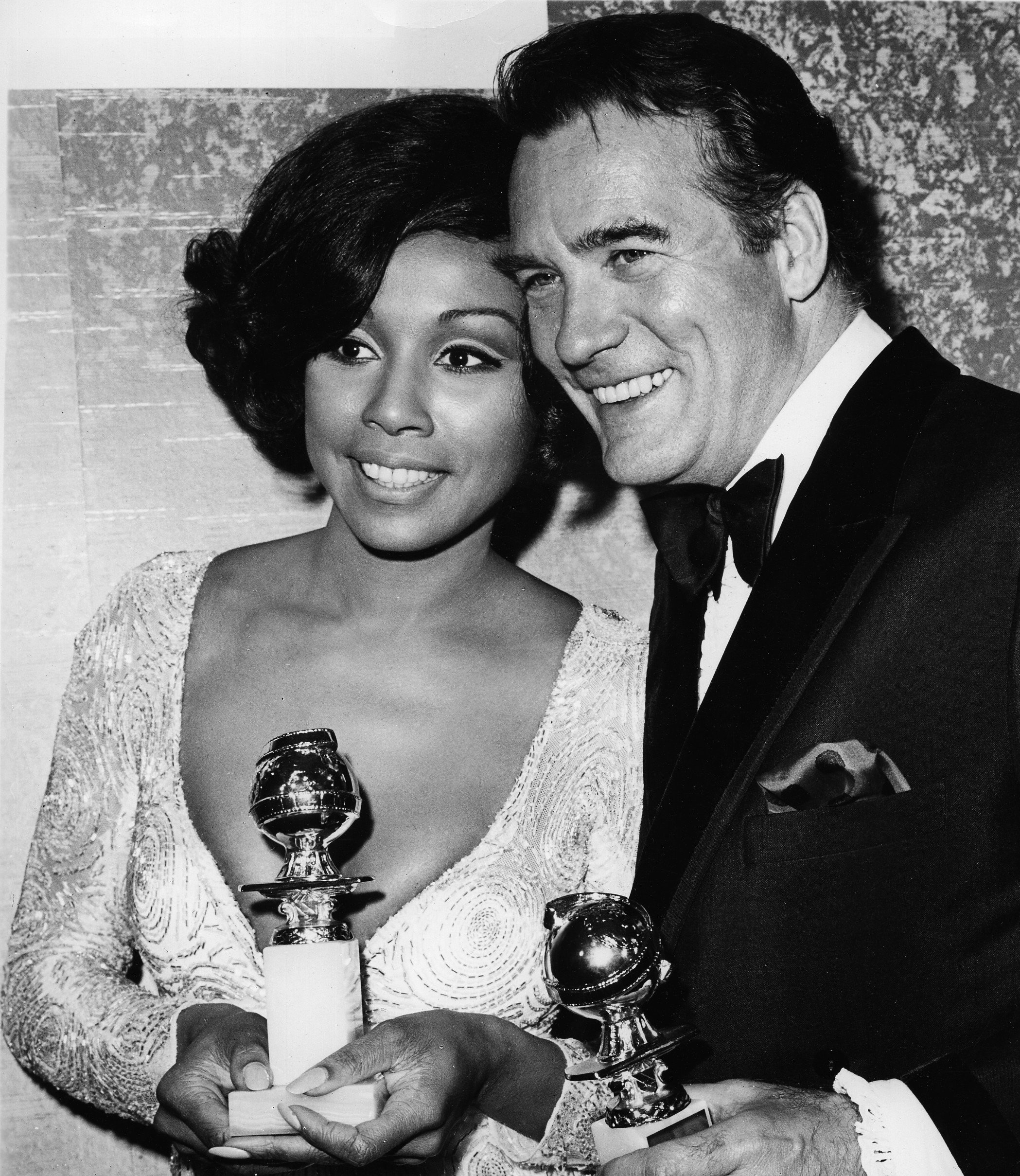 Black-and-white photo of a woman and a man at an awards ceremony, holding trophies. The woman has short black hair and is wearing a detailed, long-sleeved dress. The man has dark hair and is dressed in a tuxedo with a bow tie, smiling at the camera.