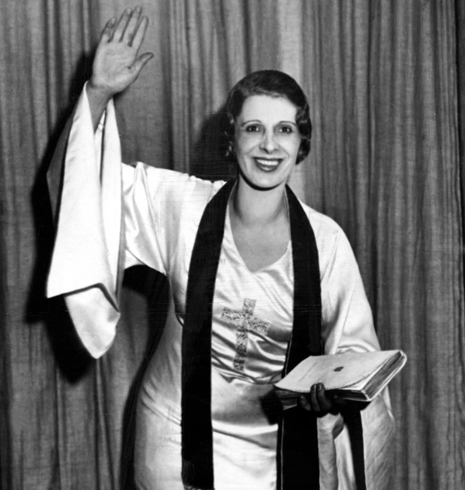 A woman in a graduation gown and stole standing against a wooden backdrop, smiling, holding a diploma, and raising her right hand.