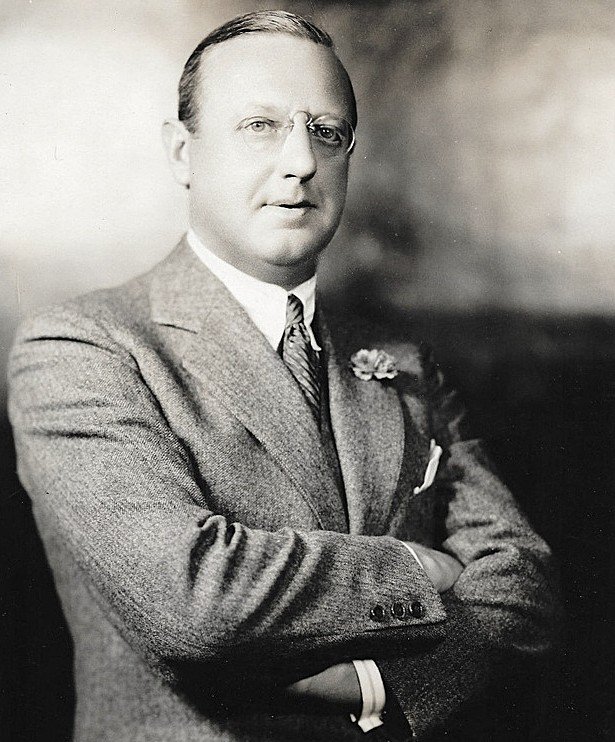 A black and white portrait of a man in a suit with glasses, arms crossed, wearing a flower on his suit lapel.