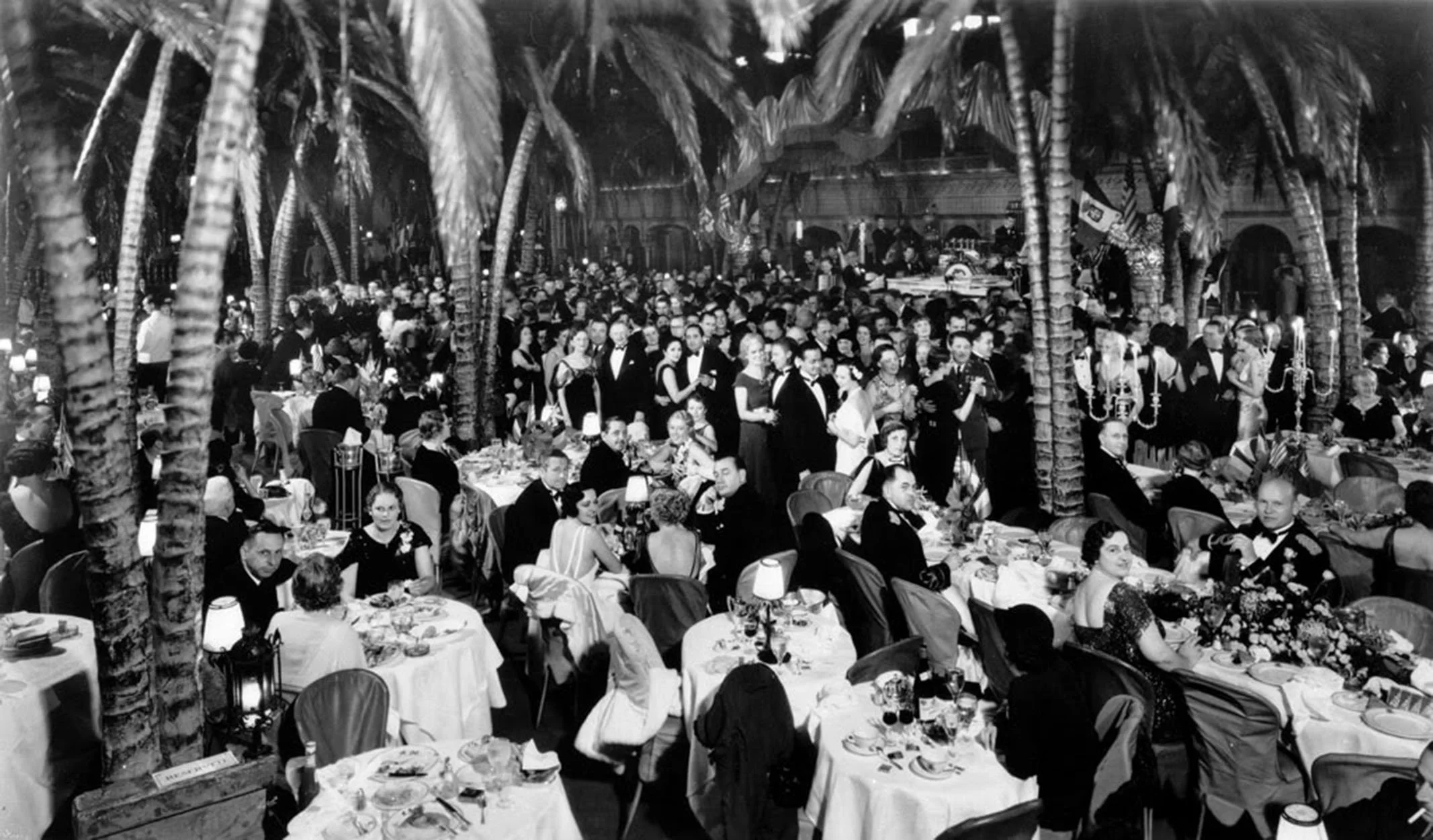 Black and white photograph of a large elegant gathering or banquet with many well-dressed people seated at tables and standing, surrounded by palm trees and decorated with table settings and candelabras, suggesting a formal event in a tropical or indoor palm-lined venue.