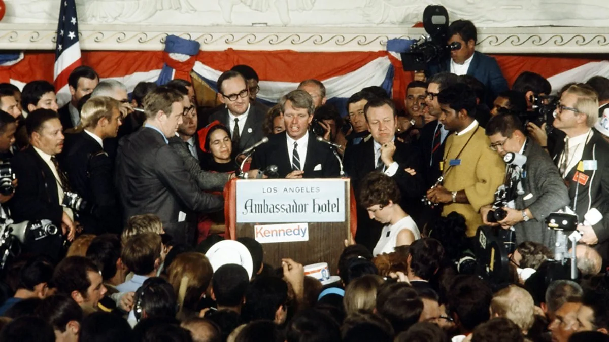 A political rally with a man speaking at a podium labeled "Los Angeles Ambassador Hotel Kennedy," surrounded by a large crowd, photographers, and press, with American flags in the background.