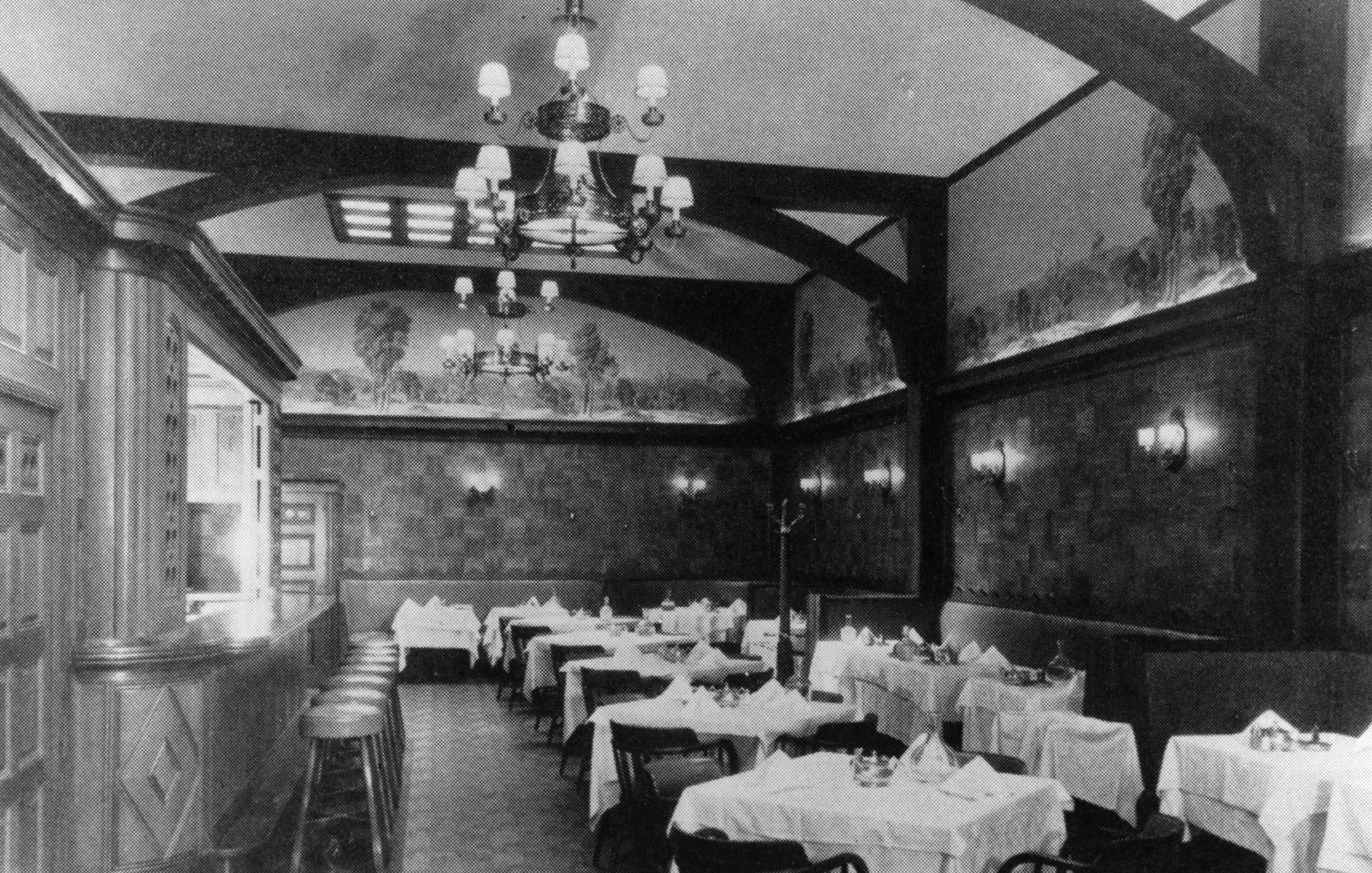 Black and white photo of an elegant restaurant interior with tables set with white tablecloths, chairs, wall sconces, chandeliers, and ornate wall murals.