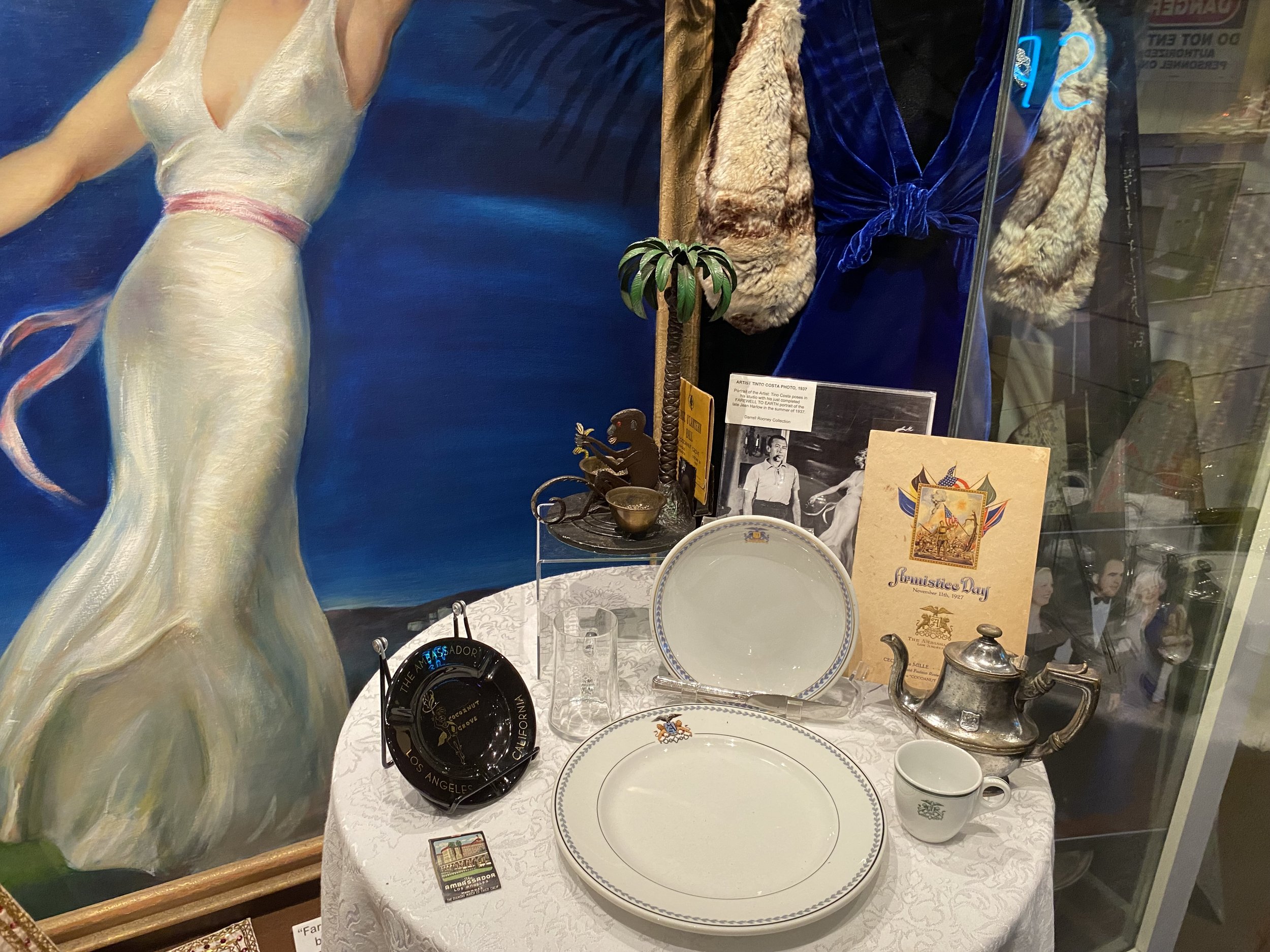 Display of vintage items in a glass case, including a large decorative plate, a teapot, a cup, vintage photographs, and memorabilia related to June 12th festival and telegraphy, with paintings and costumes visible behind.