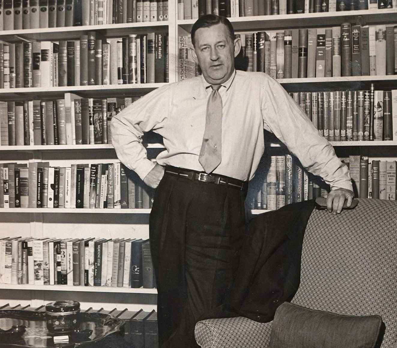 A black-and-white photo of a man in a white shirt, tie, and dark pants, standing in front of a shelf filled with books. He has one hand on his hip and the other resting on the back of a chair.