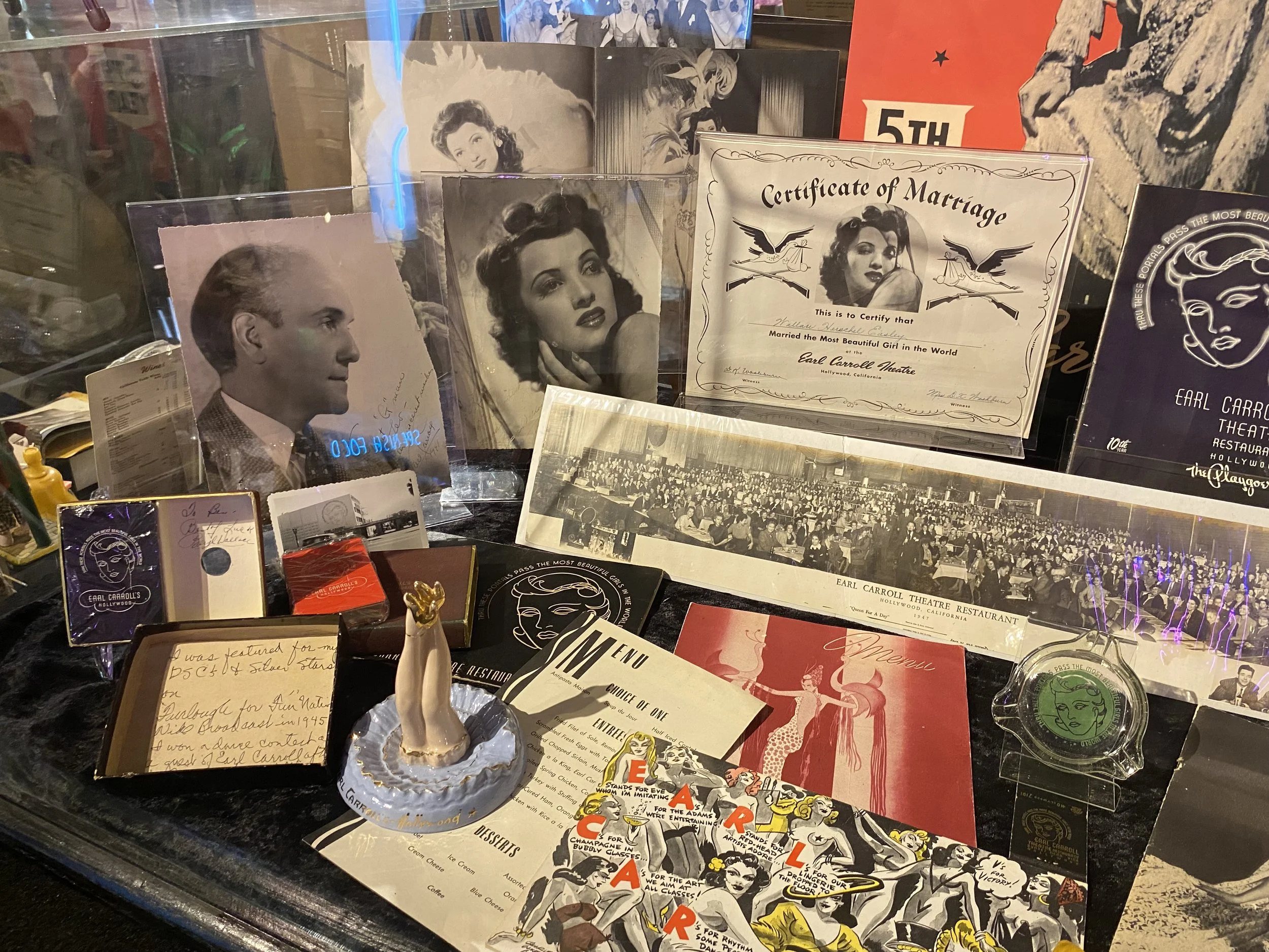 A display with vintage photographs and memorabilia related to Earl Carroll Theatre and Hollywood, including black-and-white portraits of Henry Fonda and a woman, a certificate of marriage, a large group photo of a stage or theater audience, small ite