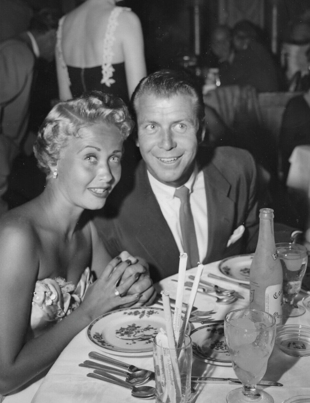 Black and white photo of a woman and man sitting at a dining table, smiling and holding hands, in a restaurant setting with other patrons in the background.