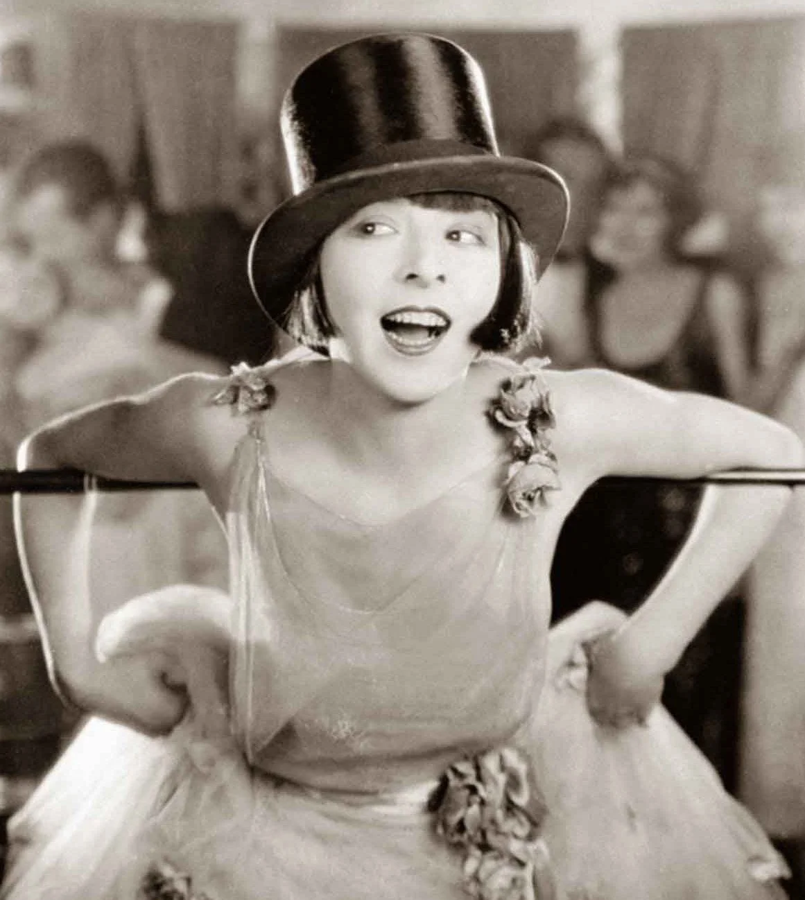 A woman wearing a large top hat and a dress with floral embellishments is smiling and appears to be in a lively setting with other people in the background.