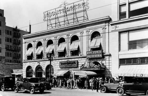 Black and white photo of a historic building with a crowd gathered outside, vintage cars parked in front, and a sign for Roy Fox's Orchestra.