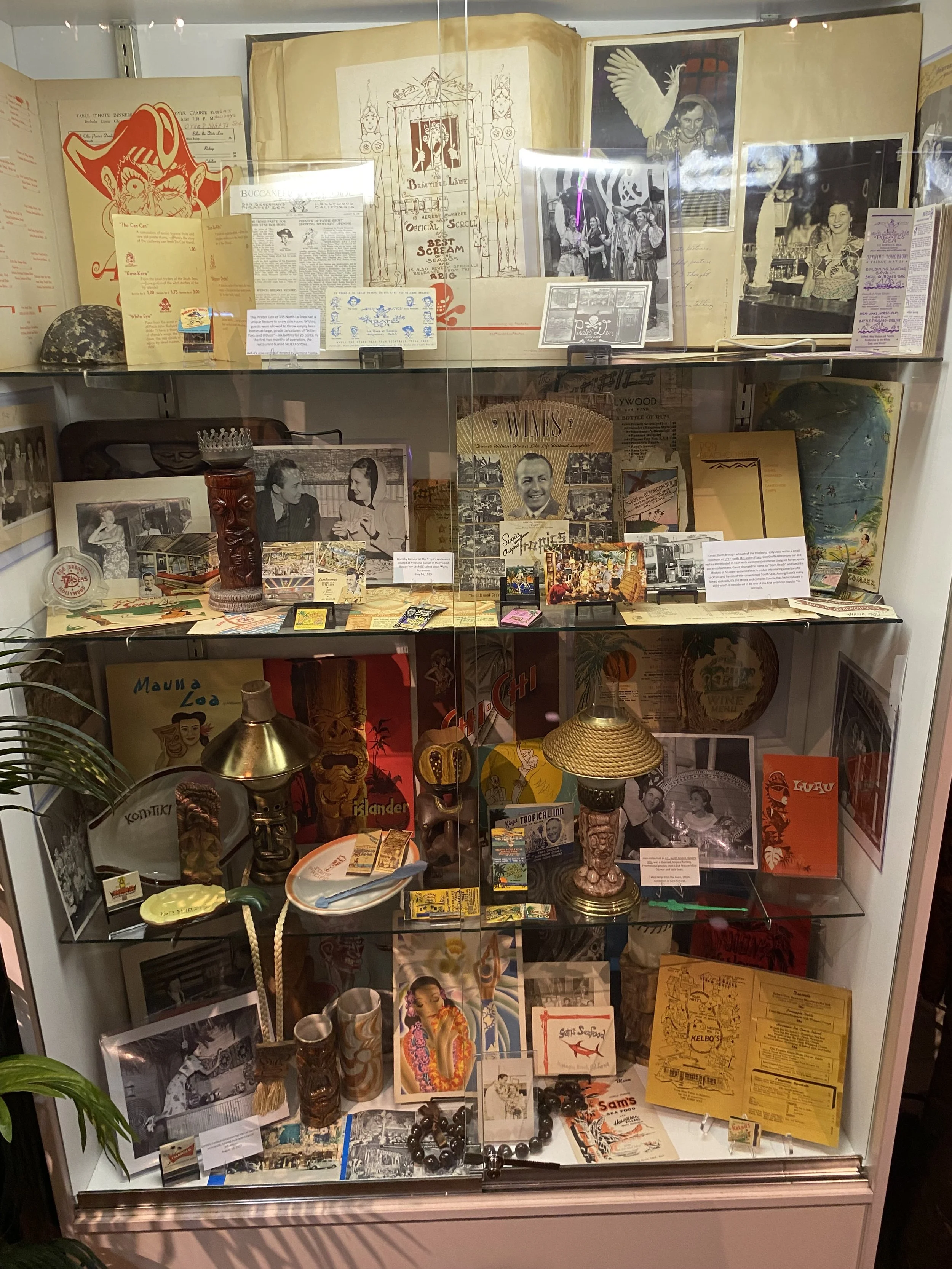 A display case filled with vintage memorabilia, black and white photographs, postcards, books, and decorative items including lamps, masks, and small souvenirs.