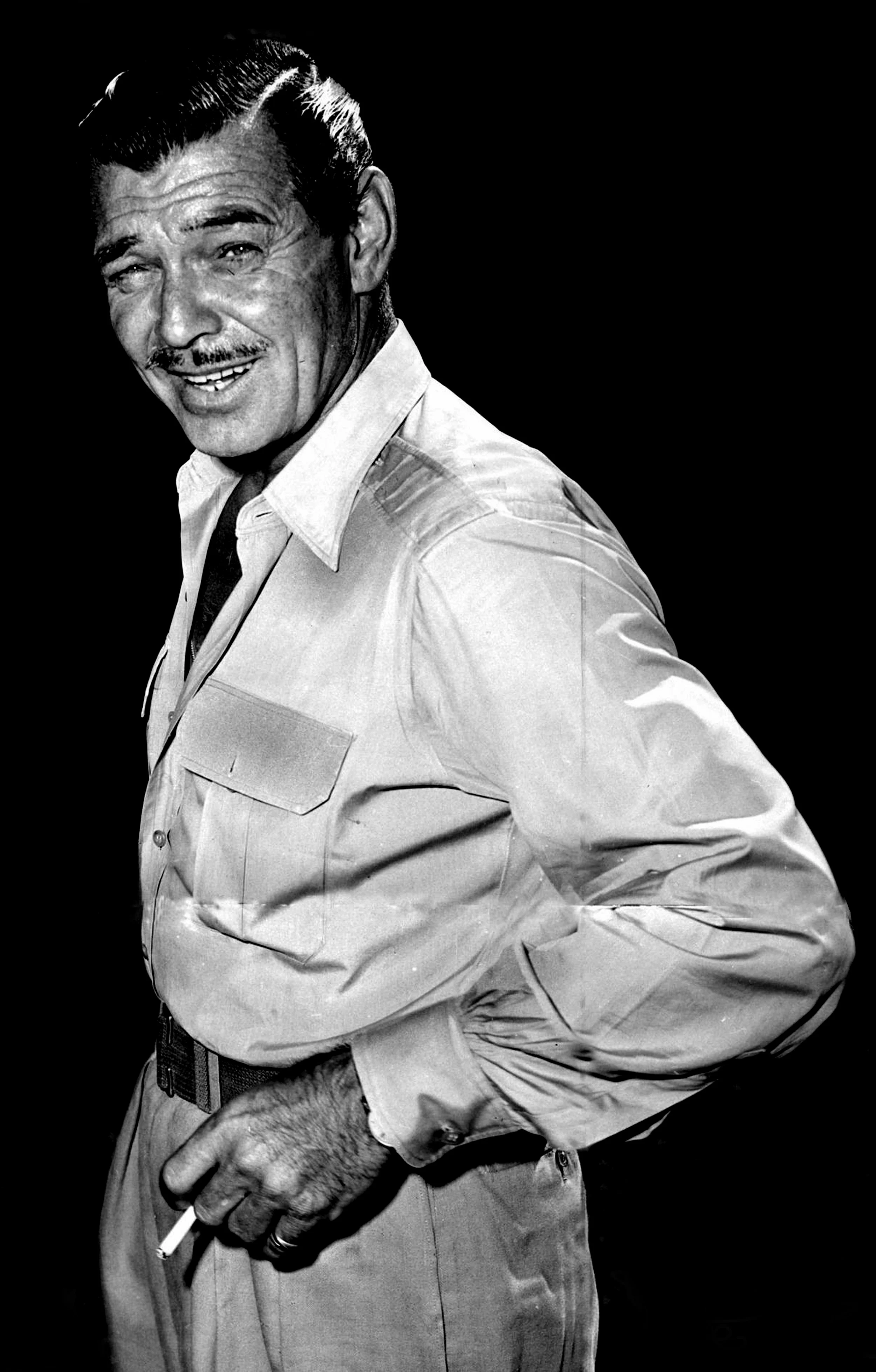 Black and white portrait of a confident man with slicked-back hair, mustache, and a cigarette in one hand, wearing a button-up shirt and trousers, smiling slightly, with a dark background.