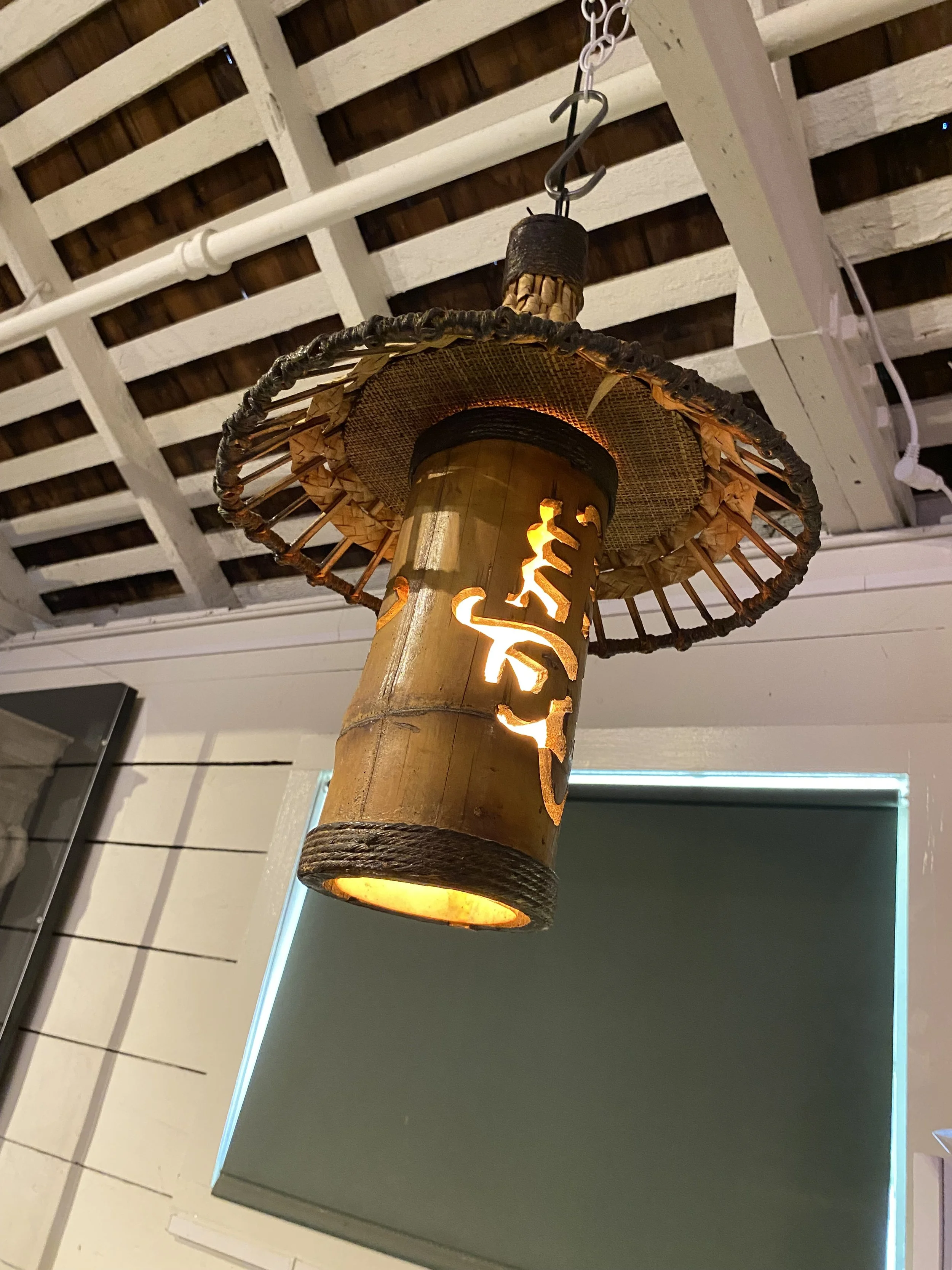 Hanging bamboo lamp with a straw hat-style shade and Japanese characters cut out, emitting warm light.