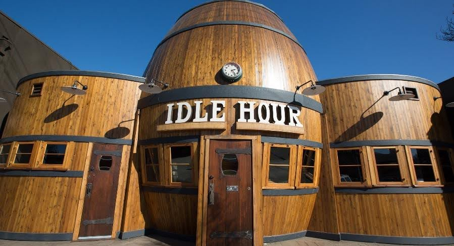 Front view of a wooden building with a rounded top, with a sign that reads 'IDLE HOUR', a clock above the sign, and three outdoor light fixtures. The building is made of wood with several windows.