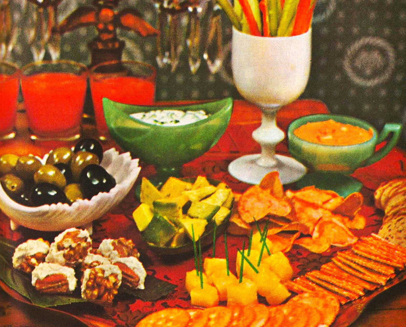 A festive table with colorful food and drinks, including a bowl of olives, several small glasses of red drink, a green bowl of salad with cucumbers, a dish of yellow dip, a bowl of soup, and various chips and crackers, with a red tablecloth and decorative background.