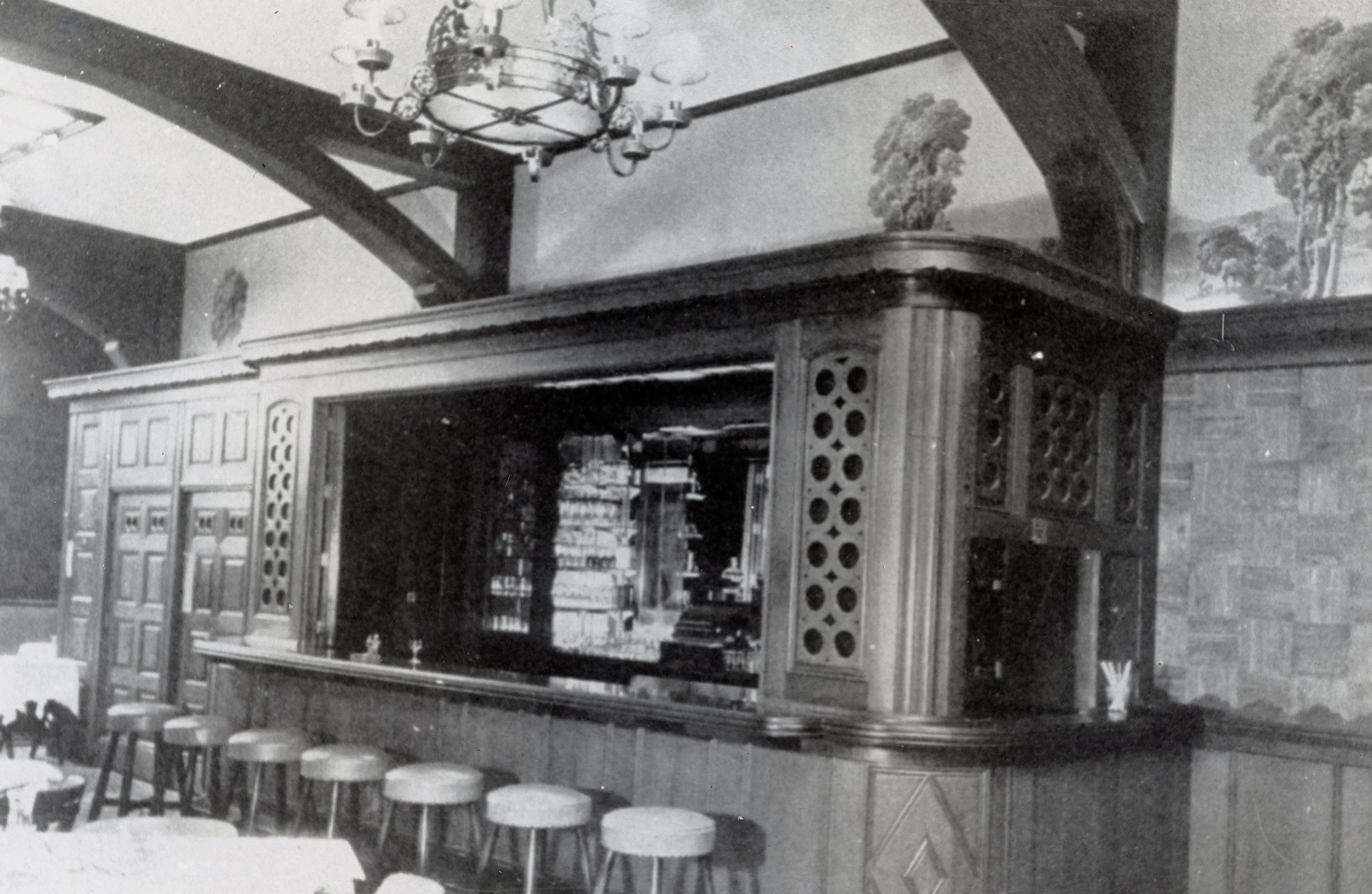 Black and white photo of a vintage bar with a wooden counter, bar stools in front, and bookshelves behind. The bar is decorated with ornate woodwork, and a chandelier hangs from the ceiling. A mural of trees is painted on the upper walls.