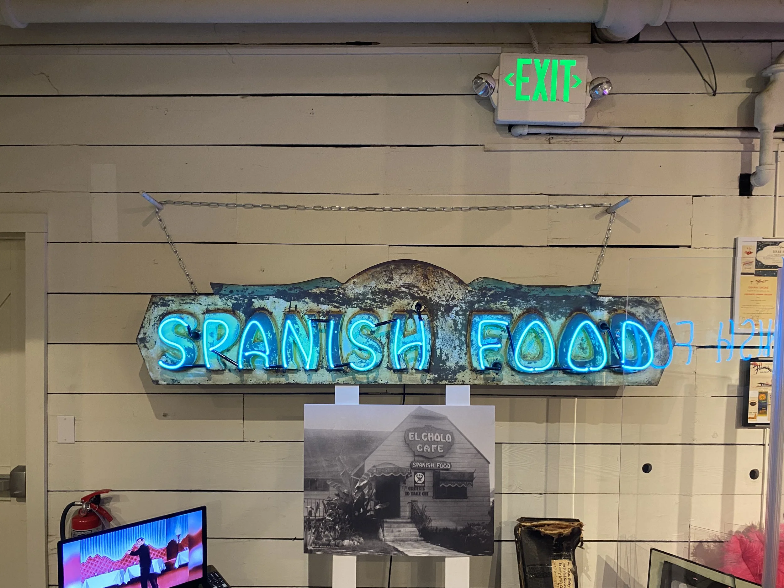 Neon sign reading 'SPANISH FOOD' hanging on a wooden wall with a green illuminated 'EXIT' sign above it.