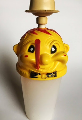 A vintage plastic soap dispenser with a smiling yellow clown face, a red nose, red spots, and a black bow tie, topped with a small beige bowl.