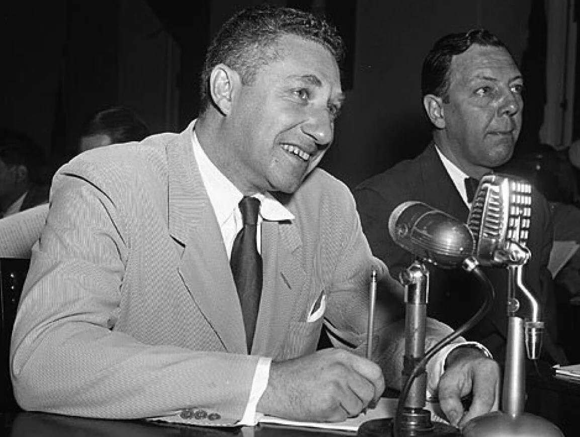 Two men dressed in suits are sitting at a table with microphones in front of them. The man in the foreground is smiling and writing notes, and the man in the background looks attentive. The image appears to be from a formal event or press conference, with a black-and-white filter.
