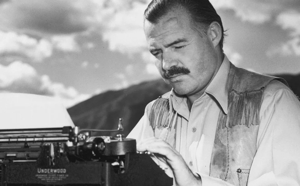A man with a mustache wearing a Western-style shirt with fringe, sitting outdoors and typing on an Underwood typewriter, with mountains and cloudy sky in the background.