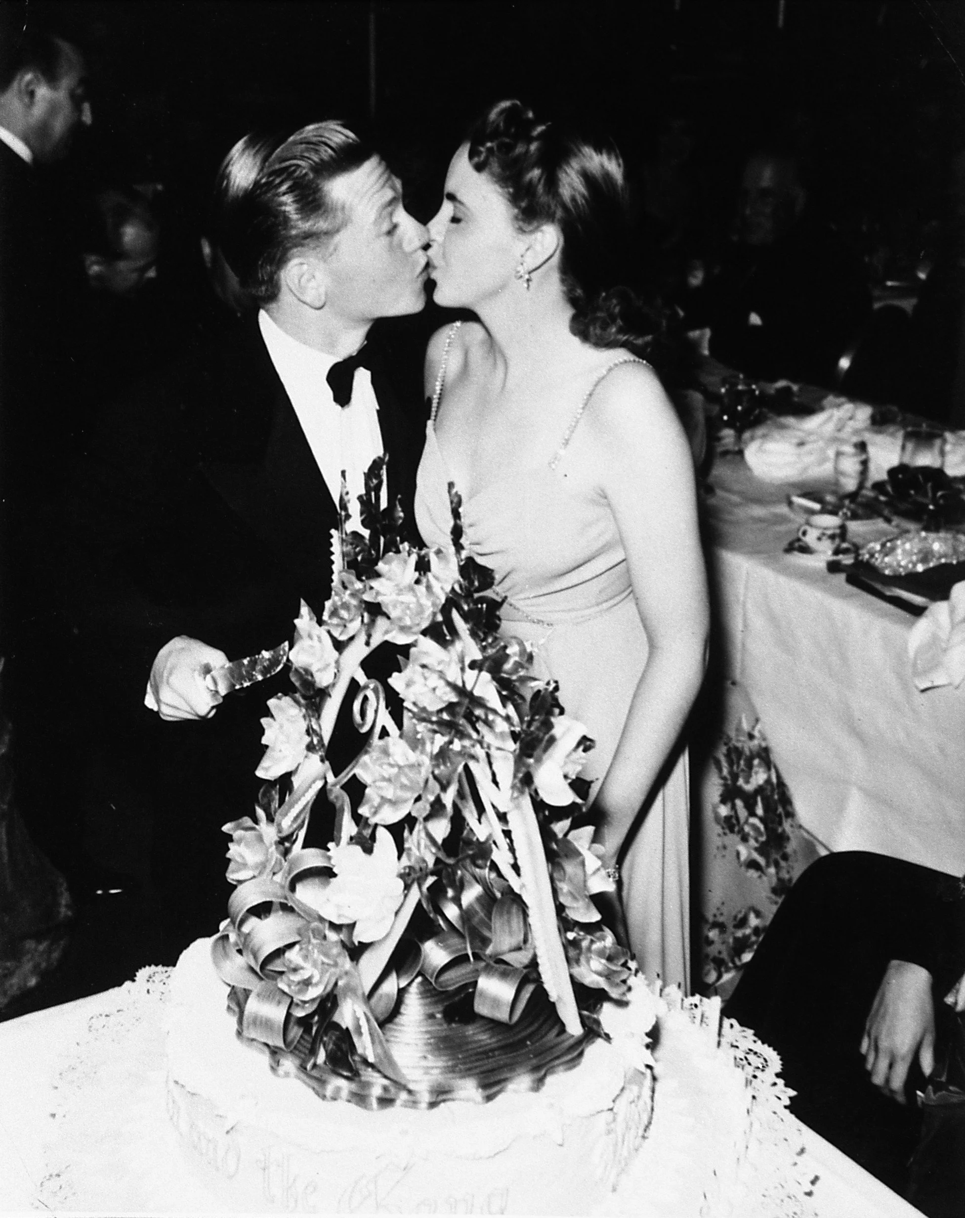 A black and white photo of a man and woman kissing at a celebration, with a large floral arrangement on a table and other people in the background.