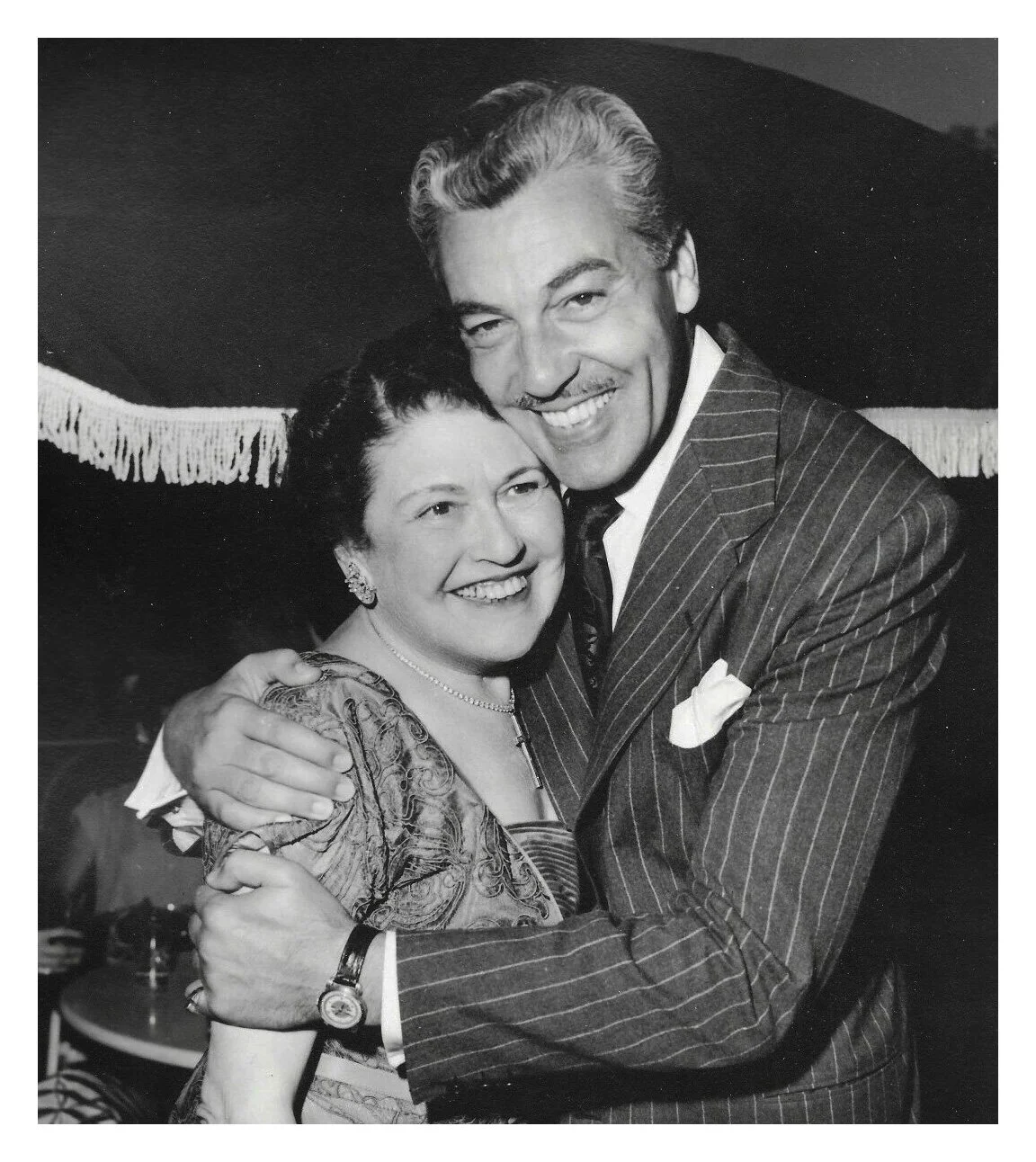 Black and white photo of a smiling man and woman hugging, dressed in vintage attire, likely from the mid-20th century.