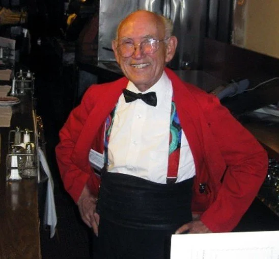 An elderly man with glasses, wearing a red jacket, white shirt, black bow tie, and suspenders, smiling at a restaurant.