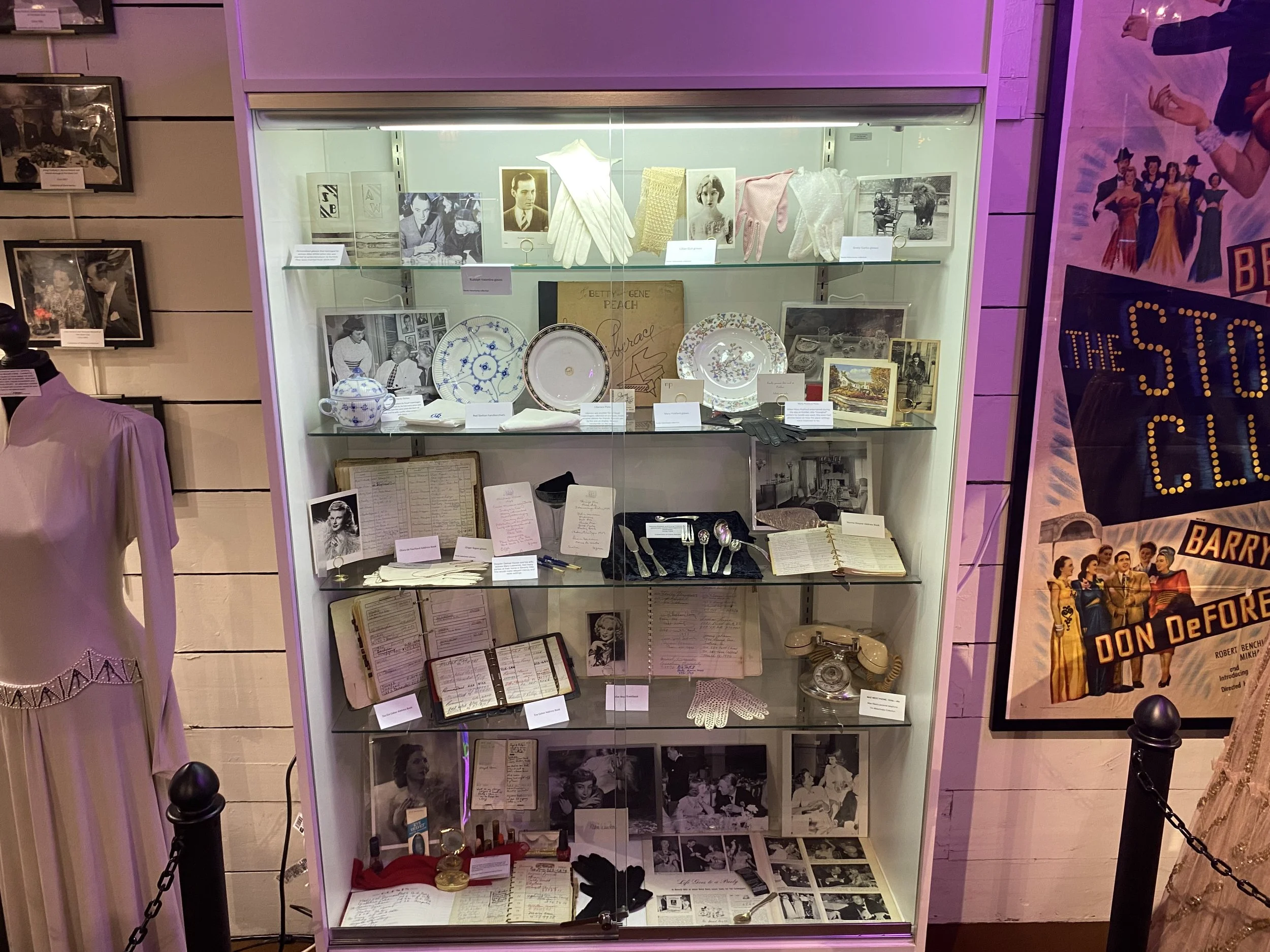 A display case containing vintage memorabilia, including photographs, plates, books, gloves, and other collectible items, with a mannequin in a pink dress to the left and a poster to the right.