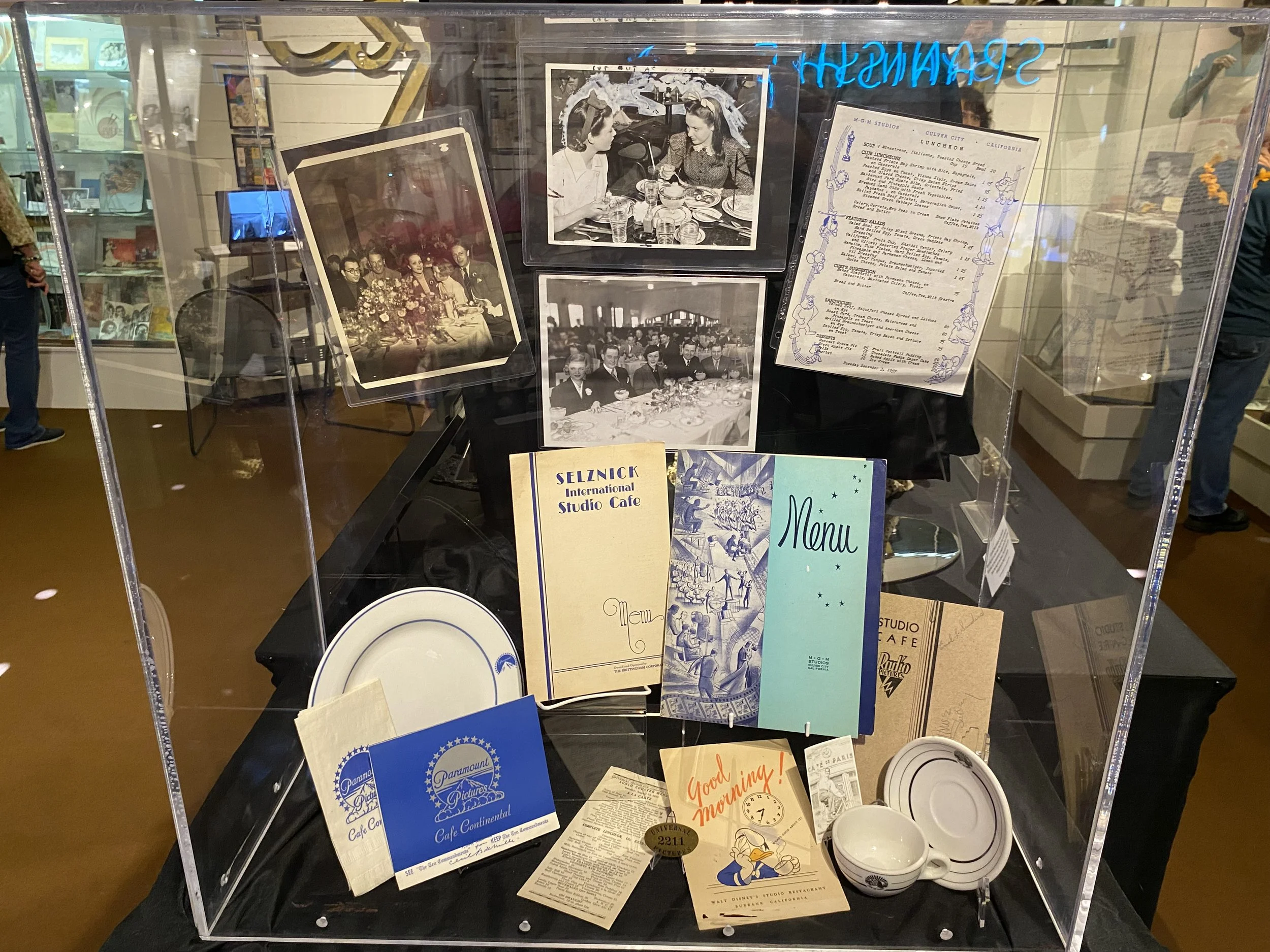 Display case with black and white photographs, menus, napkins, paper plates, and a bowl, commemorating a historic cafe from Paramount Pictures, with reflections of the museum interior in the glass.