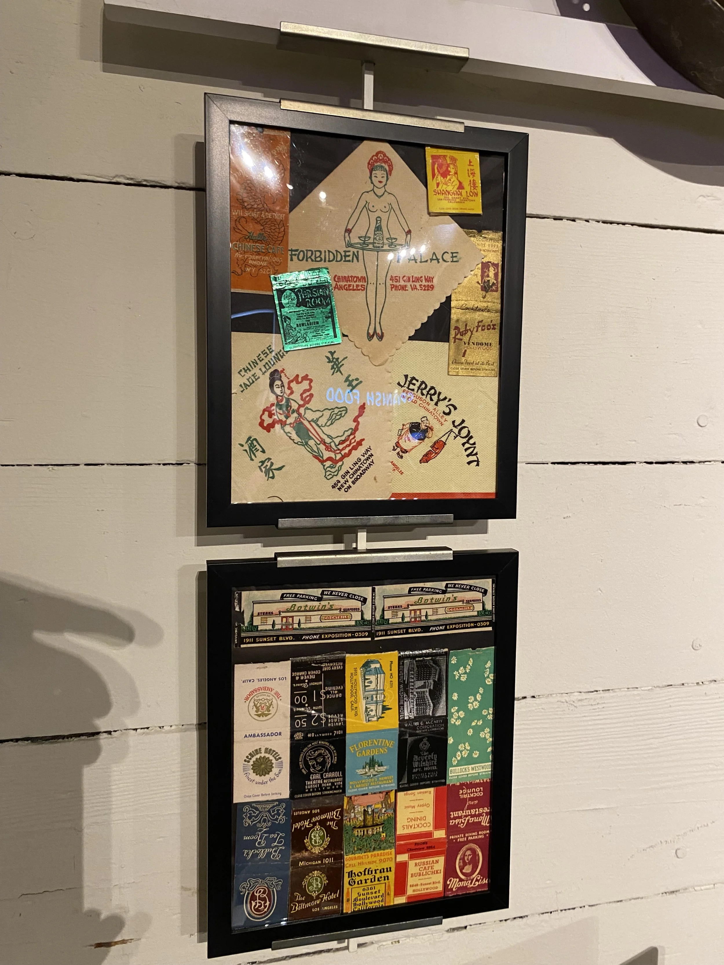 Two framed collages with vintage-style paper items such as tickets, stickers, drawings, and advertisements, displayed on a white wooden wall.