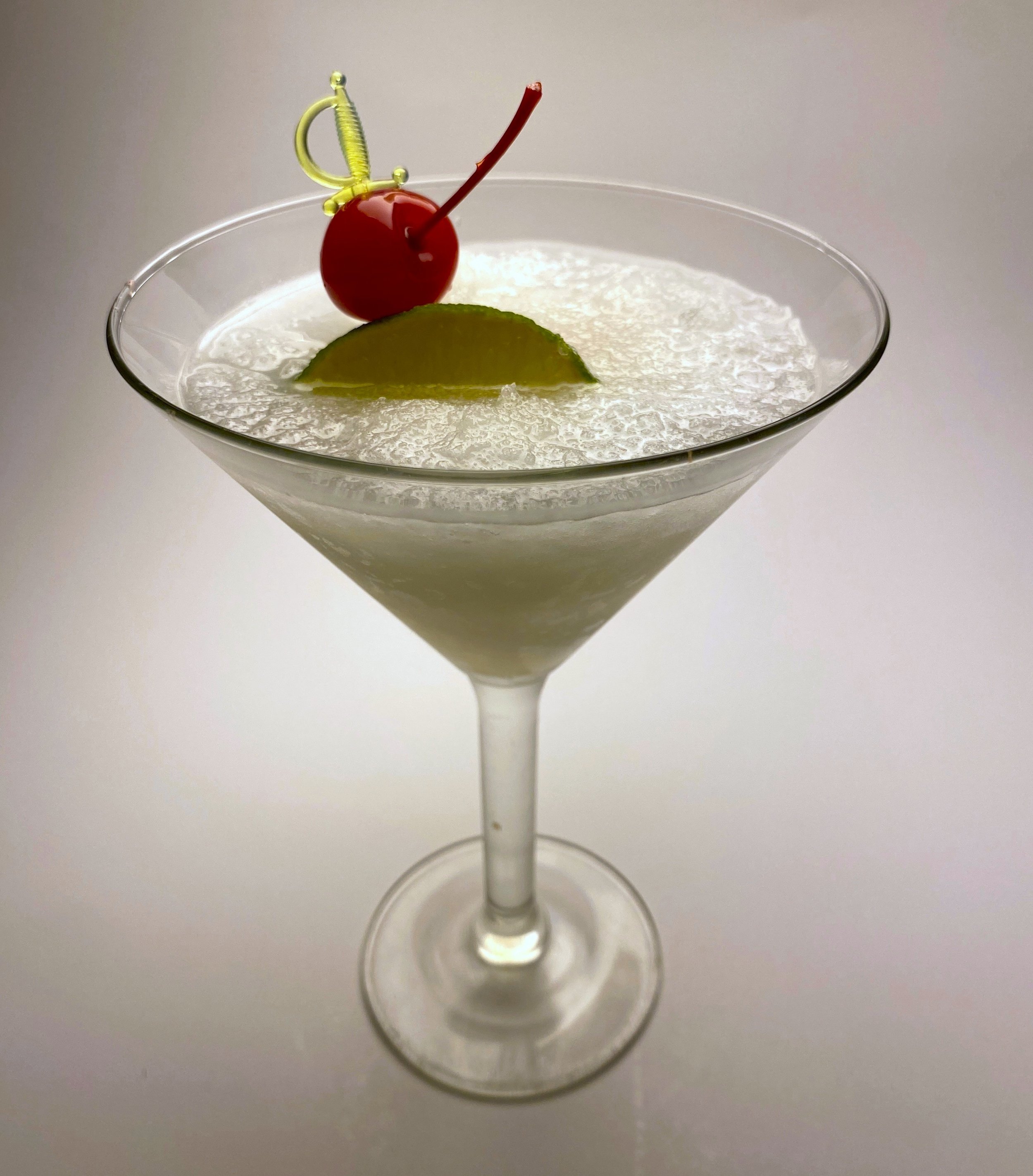 A classic martini glass filled with a creamy white cocktail, garnished with a maraschino cherry, a lime wedge, and a decorative cocktail pick.
