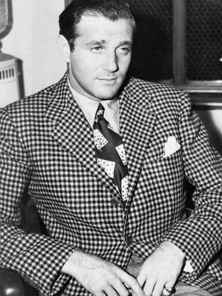 Black-and-white photo of a man wearing a houndstooth patterned suit and a shirt with a geometric design, sitting with hands clasped in front of him, in front of a textured background.