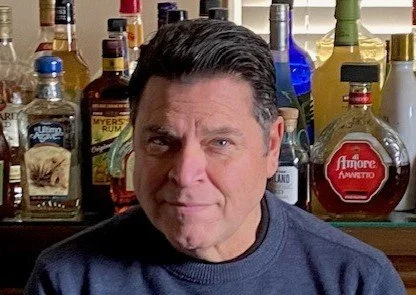 A man with dark hair making a skeptical facial expression, standing in front of a bar with various bottles of alcohol.