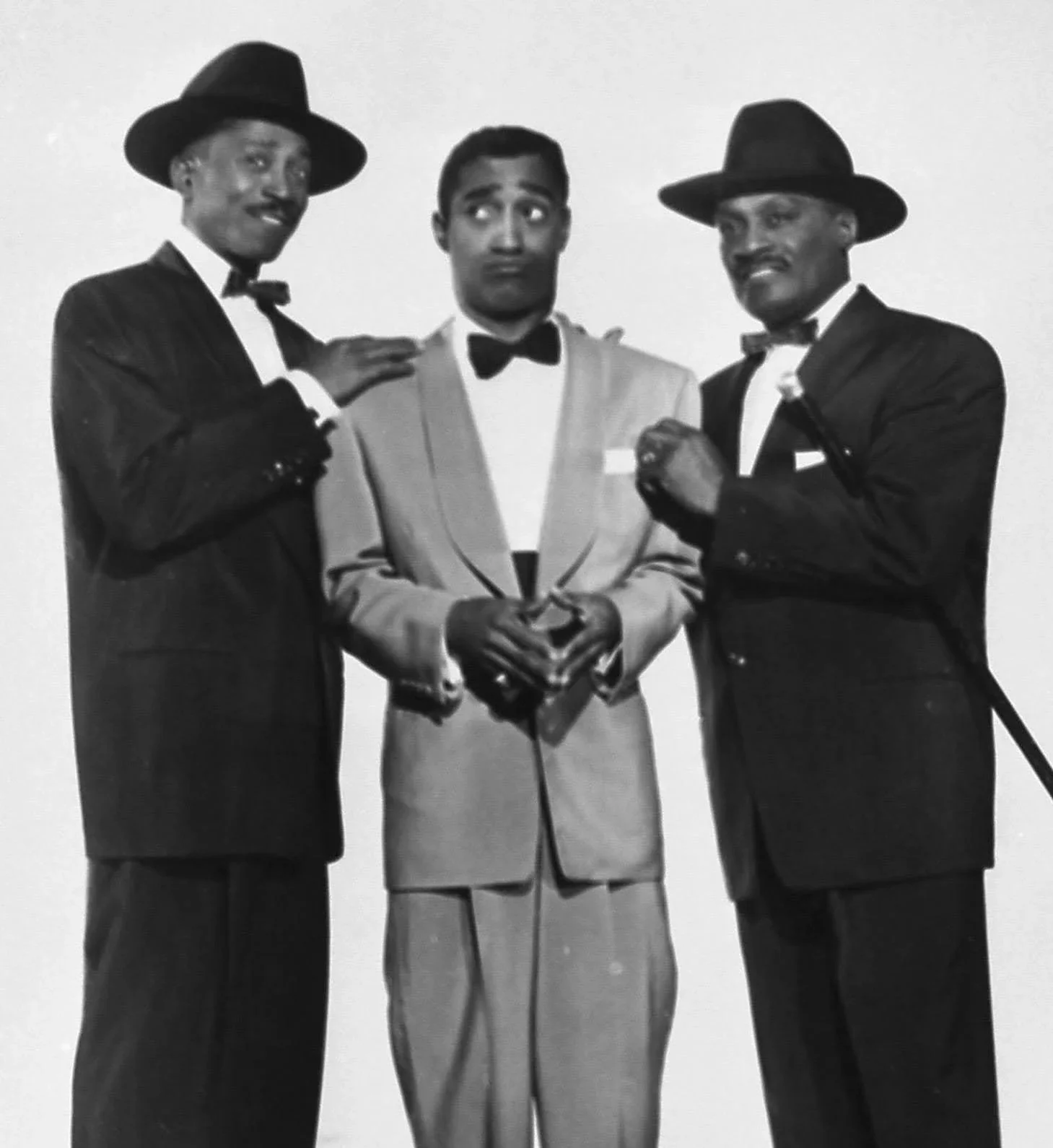 Black and white photo of three men dressed in formal tuxedos and hats, posing together. The man in the middle has a surprised or confused expression, while the men on either side are smiling.
