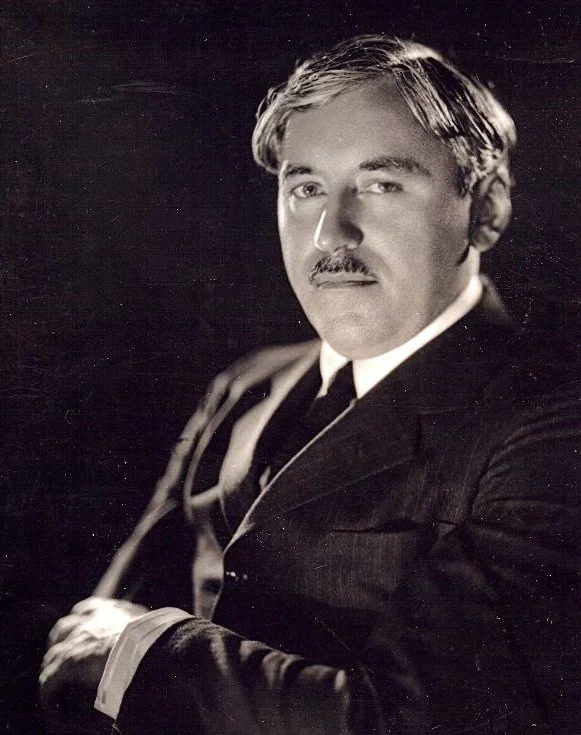 Black and white portrait of a man with light-colored hair and a mustache, wearing a dark suit and tie, looking slightly to the camera with a serious expression, seated with his arms crossed.