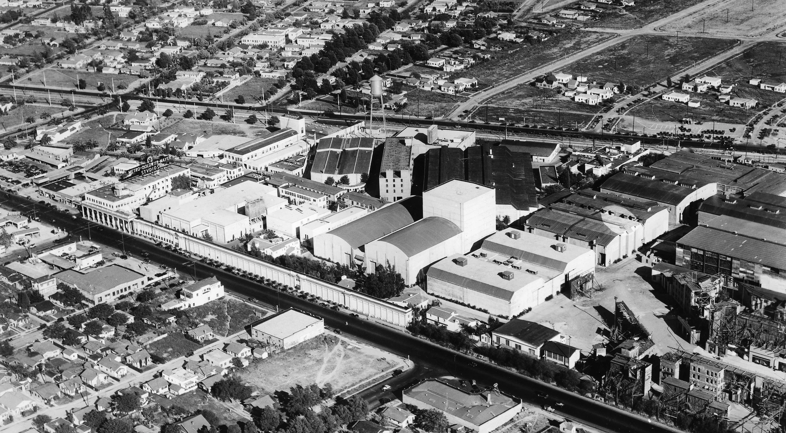 Aerial black and white photograph of a cityscape showing industrial warehouses, commercial buildings, residential houses, and streets. A water tower is visible among the buildings, and train tracks run through the area.
