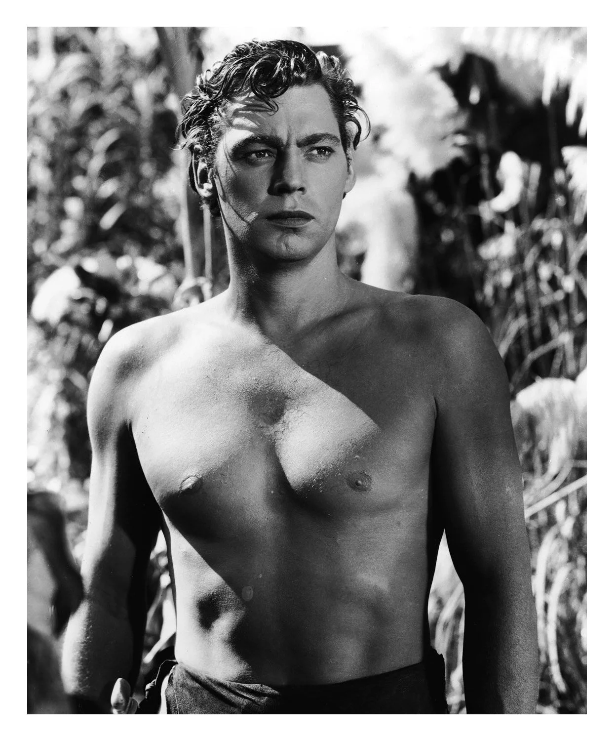 A black and white photo of a shirtless man with wavy hair standing outdoors with blurred foliage in the background.