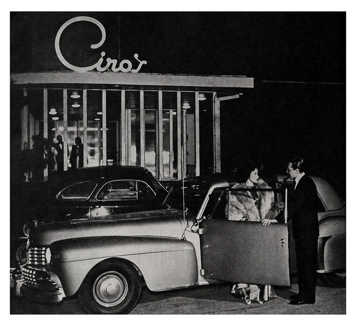 Night scene outside a drive-in restaurant with a vintage car in the foreground. Two people, a man and a woman, are standing beside the car, engaged in conversation. Other people are visible inside the restaurant. The restaurant sign reads "Ciro's".