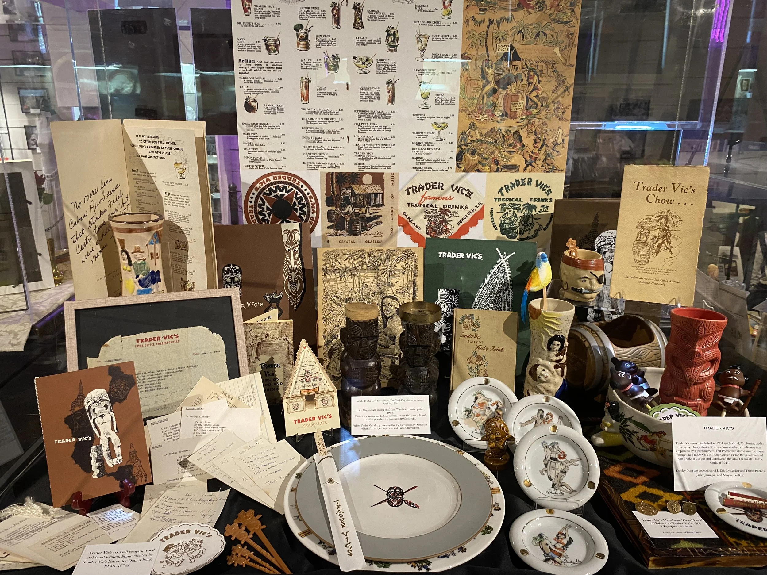 A display window with various Trader Vic's memorabilia including menus, artwork, souvenirs, ceramics, and small figurines. There are tiki mugs, decorative plates, carved statues, and printed materials related to Trader Vic's history and tropical drin