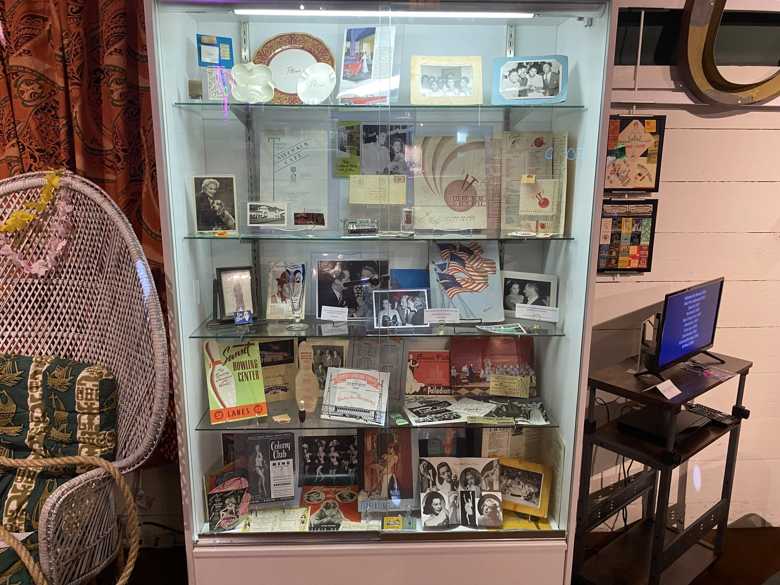 A glass display case filled with vintage photographs, posters, memorabilia, and printed materials, situated next to a wicker chair with patterned cushions. To the right, a small black table with a monitor screen.