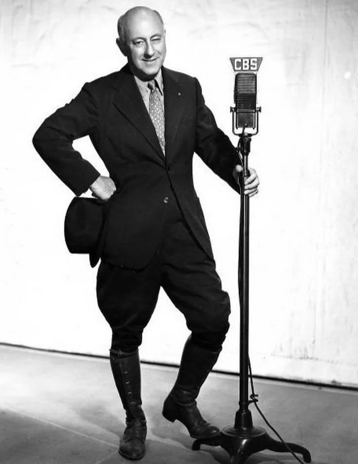 A man standing next to a CBS microphone, dressed in a suit, holding a hat, with his other hand on his hip, in a black-and-white photo.