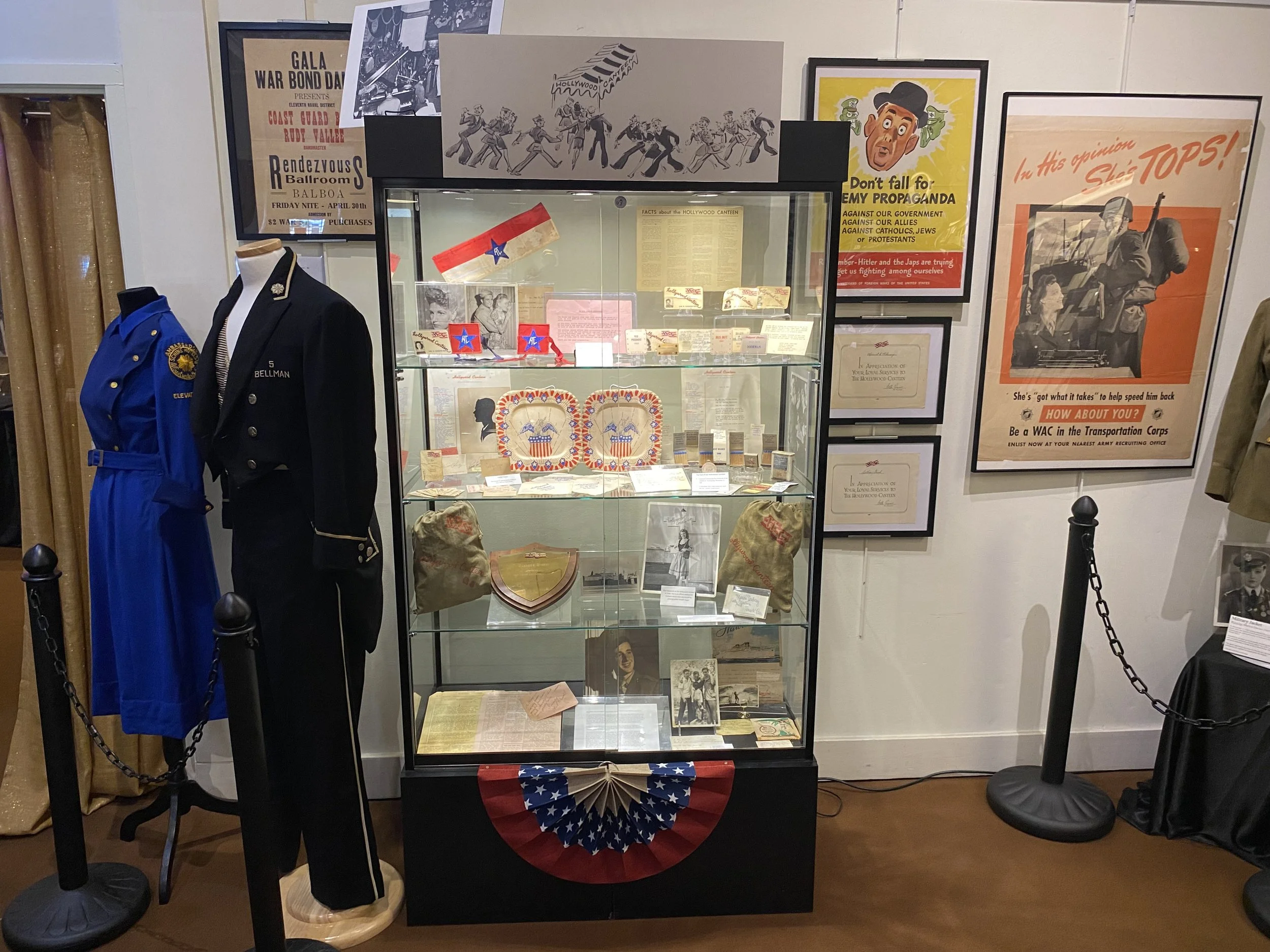 Exhibit display of historical items including military uniforms, photographs, and memorabilia, surrounded by posters advocating war bond donations and remembrance, in a museum or memorial setting.