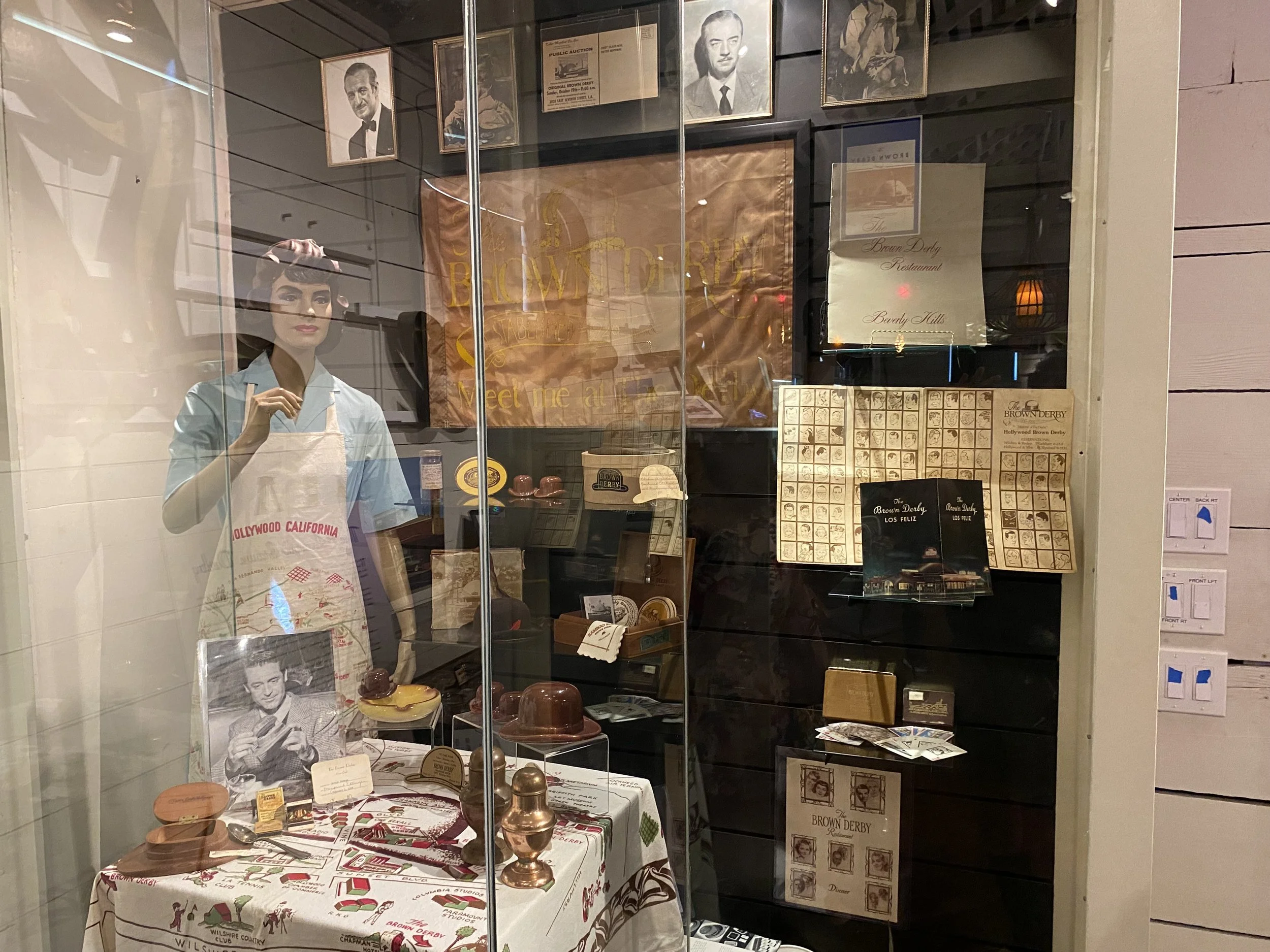 A vintage display window featuring a mannequin dressed as a waitress, photographs of classic actors, and various memorabilia including a table with a tablecloth, a framed photo of a man, and boxes of chocolates. Behind the glass, there are signs and 