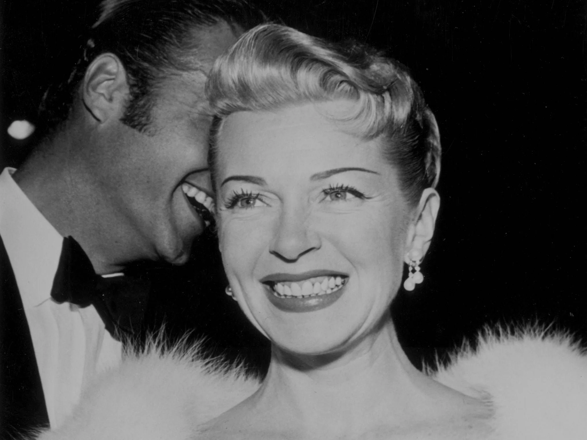 Black and white photo of a man whispering in a woman's ear, both smiling. Woman has short light-colored hair, earrings, and a fluffy dress or shawl.