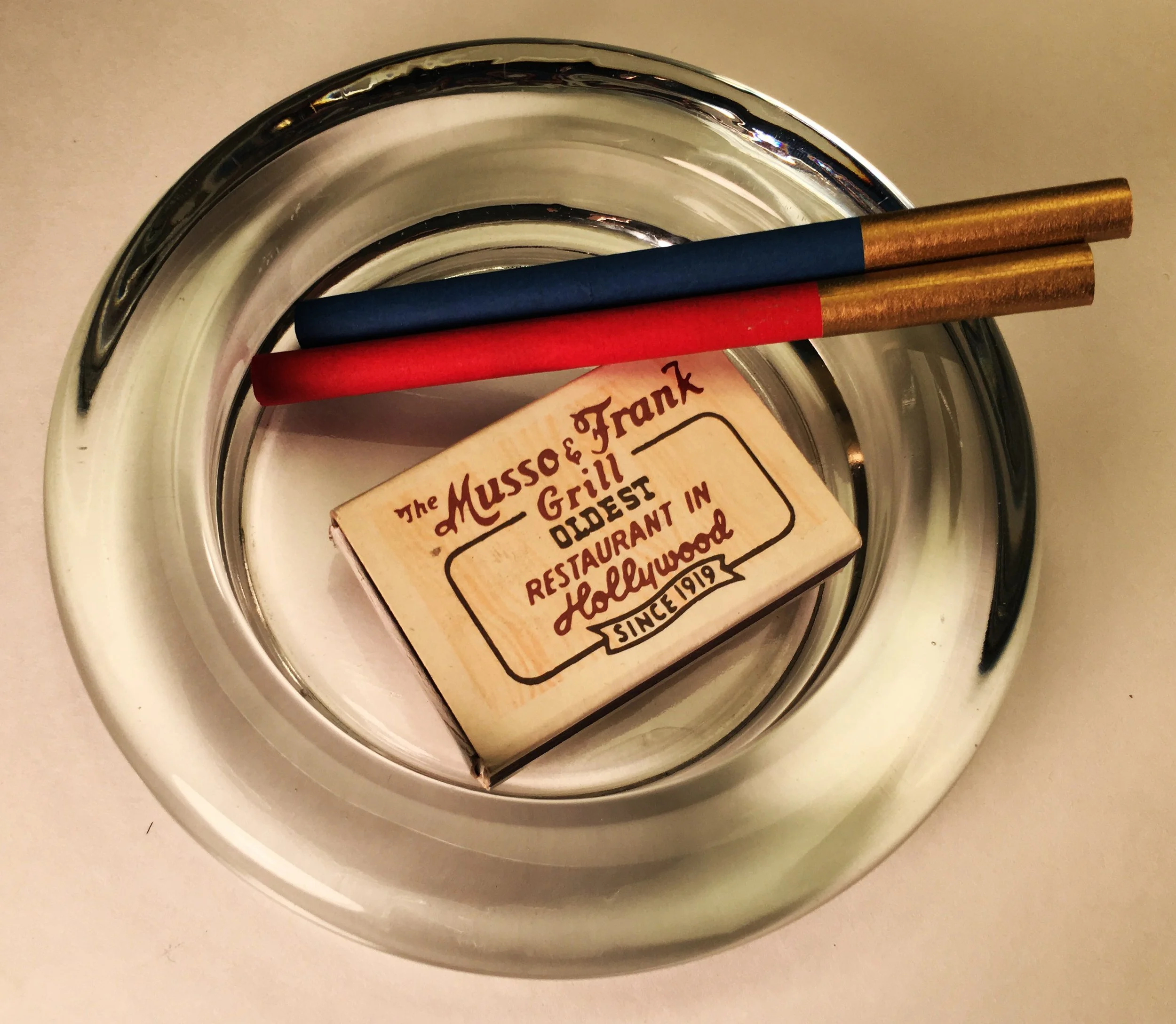 A metal ashtray containing a wooden matchbook marked 'The Musso & Frank Grill, Oldest Restaurant in Hollywood, Since 1919' and two matches with gold and red tips.