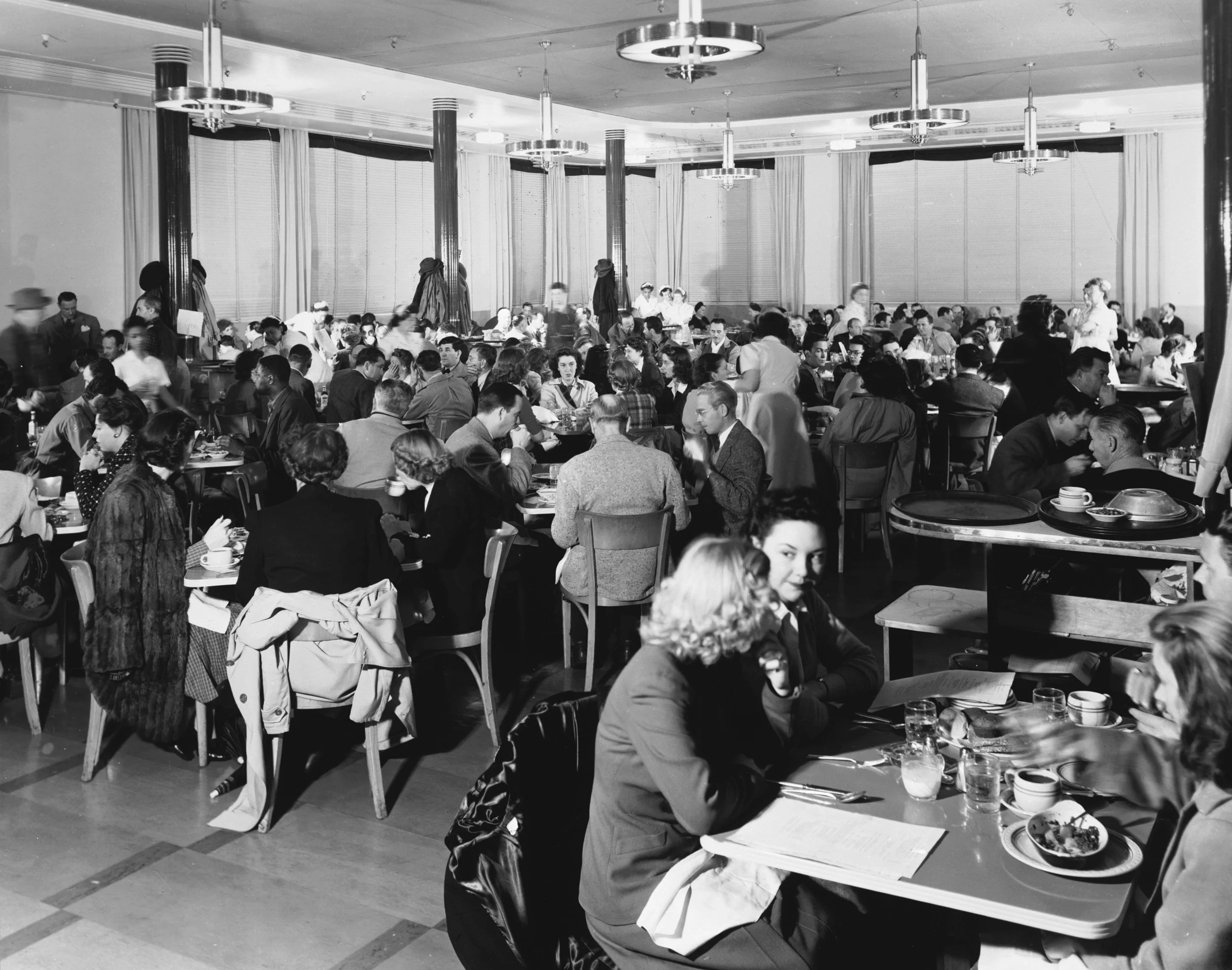 A busy indoor restaurant or cafeteria filled with people dining at tables, with some servers walking around; vintage black-and-white photo.