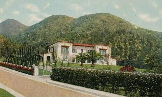 Large luxurious house with white walls, red-tiled roof, surrounded by palm trees and a well-manicured hedge, set against green mountains in the background.