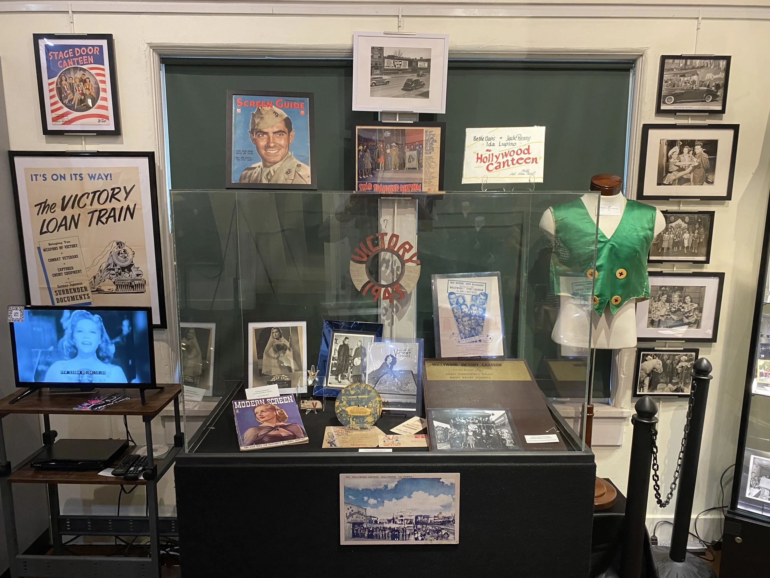 Historical display in a museum dedicated to Hollywood Canteen with photographs, posters, and memorabilia relating to World War II, including a mannequin wearing a green vest, framed images, and a glass case.