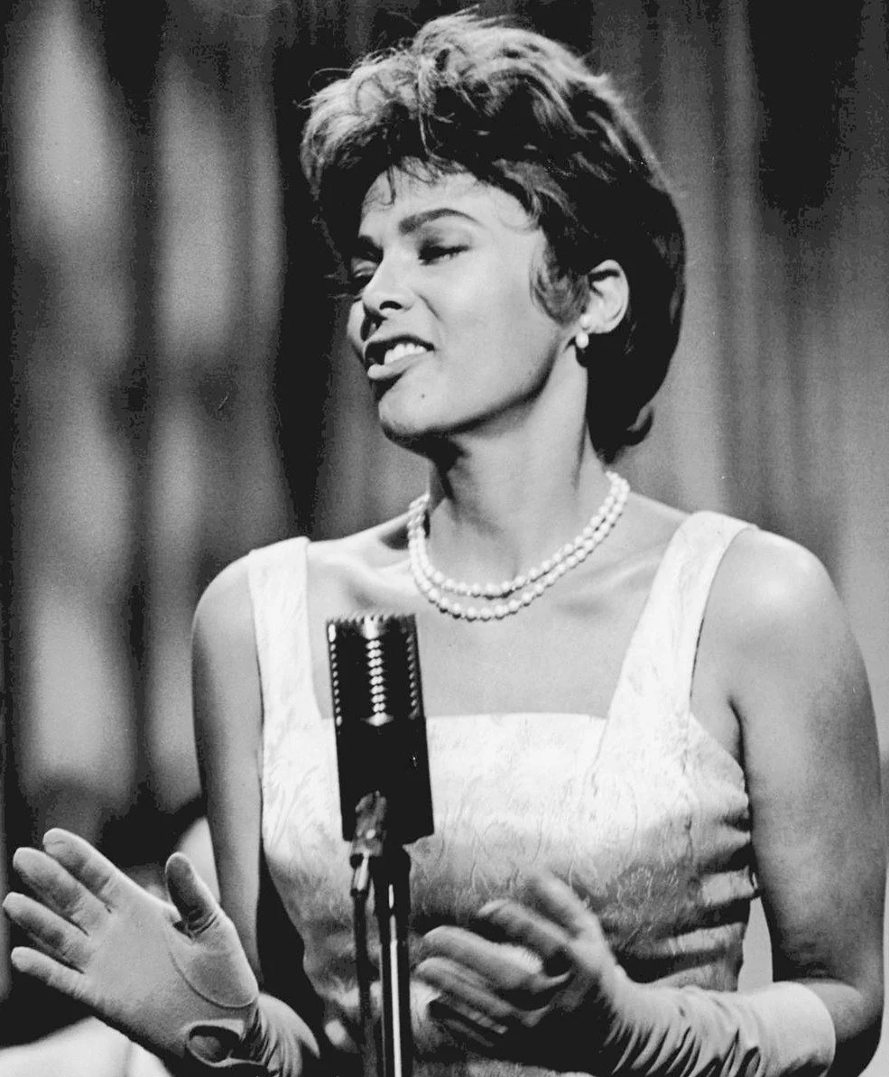 Black and white photo of a woman speaking into a vintage microphone, wearing a sleeveless dress and pearl necklace, with short styled hair, displaying a playful expression.